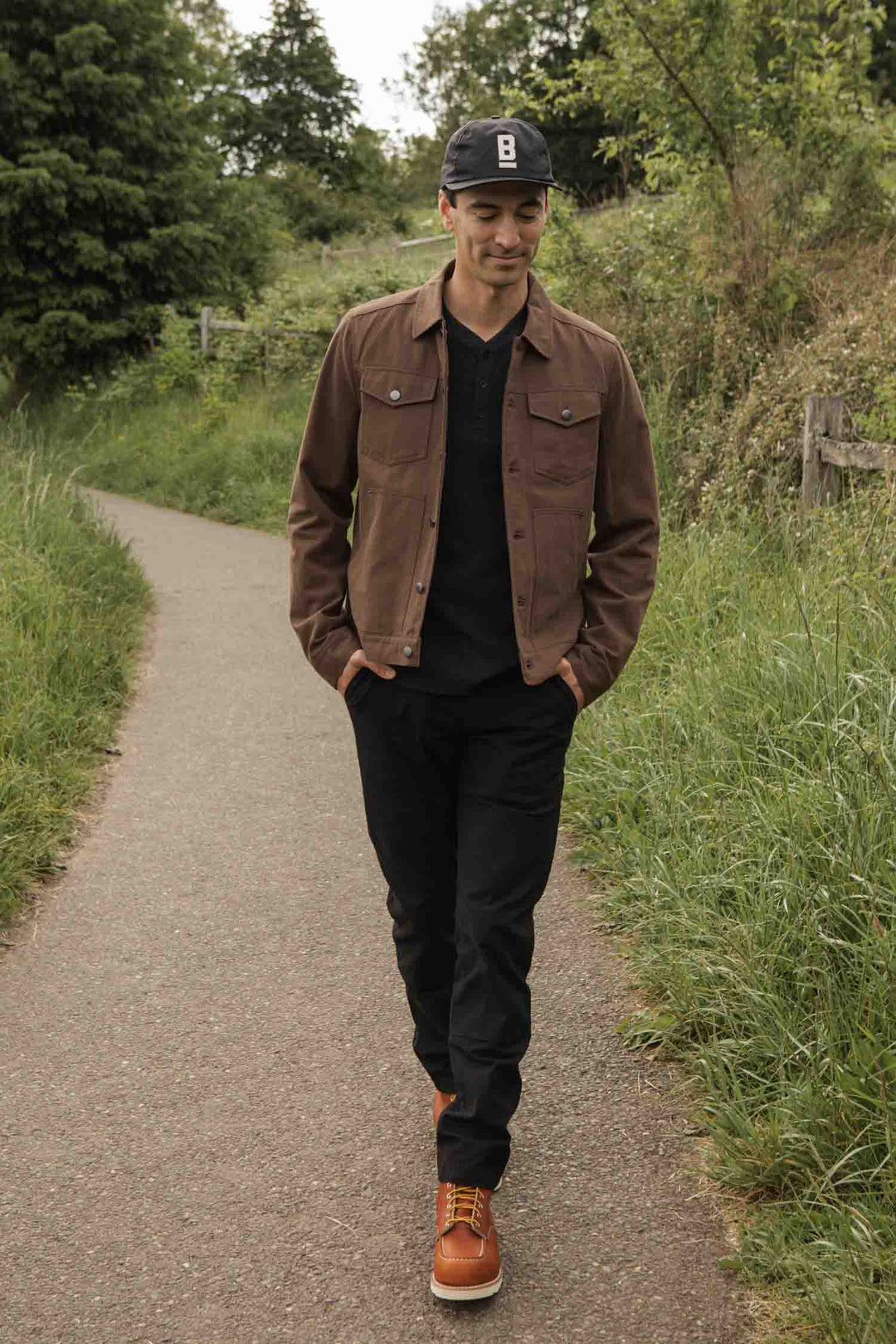 A man in the Bridge & Burn Burl Trucker Jacket in Walnut, black shirt and pants, orange boots, and a black cap walks along a paved path through greenery with his hands in his pockets and a slight smile.