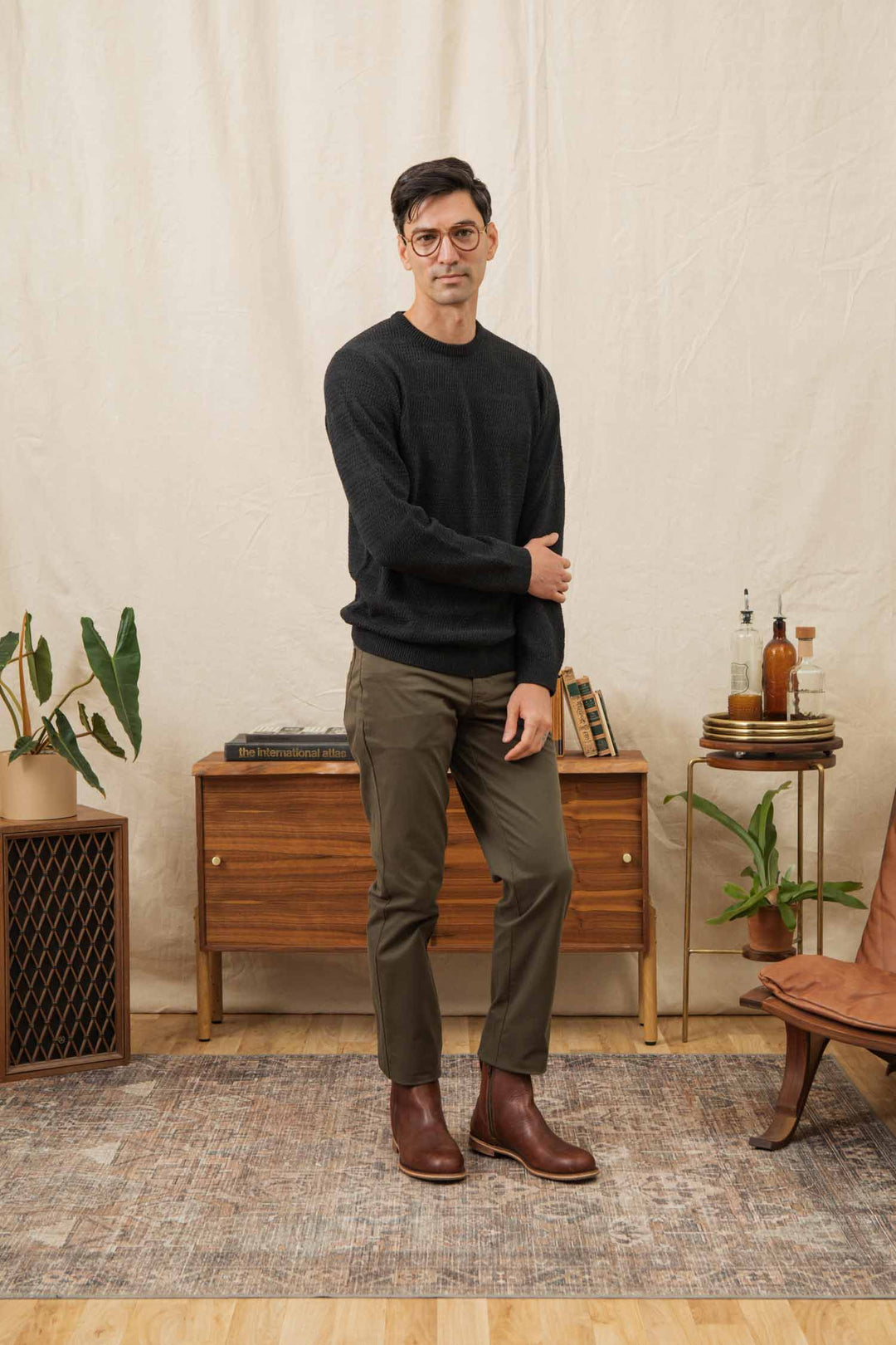 A man with short dark hair and glasses wears the Bridge & Burn Calder Sweater in Midnight, olive pants, and brown boots, standing in a mid-century room filled with plants, books, and wooden furniture atop a patterned rug.