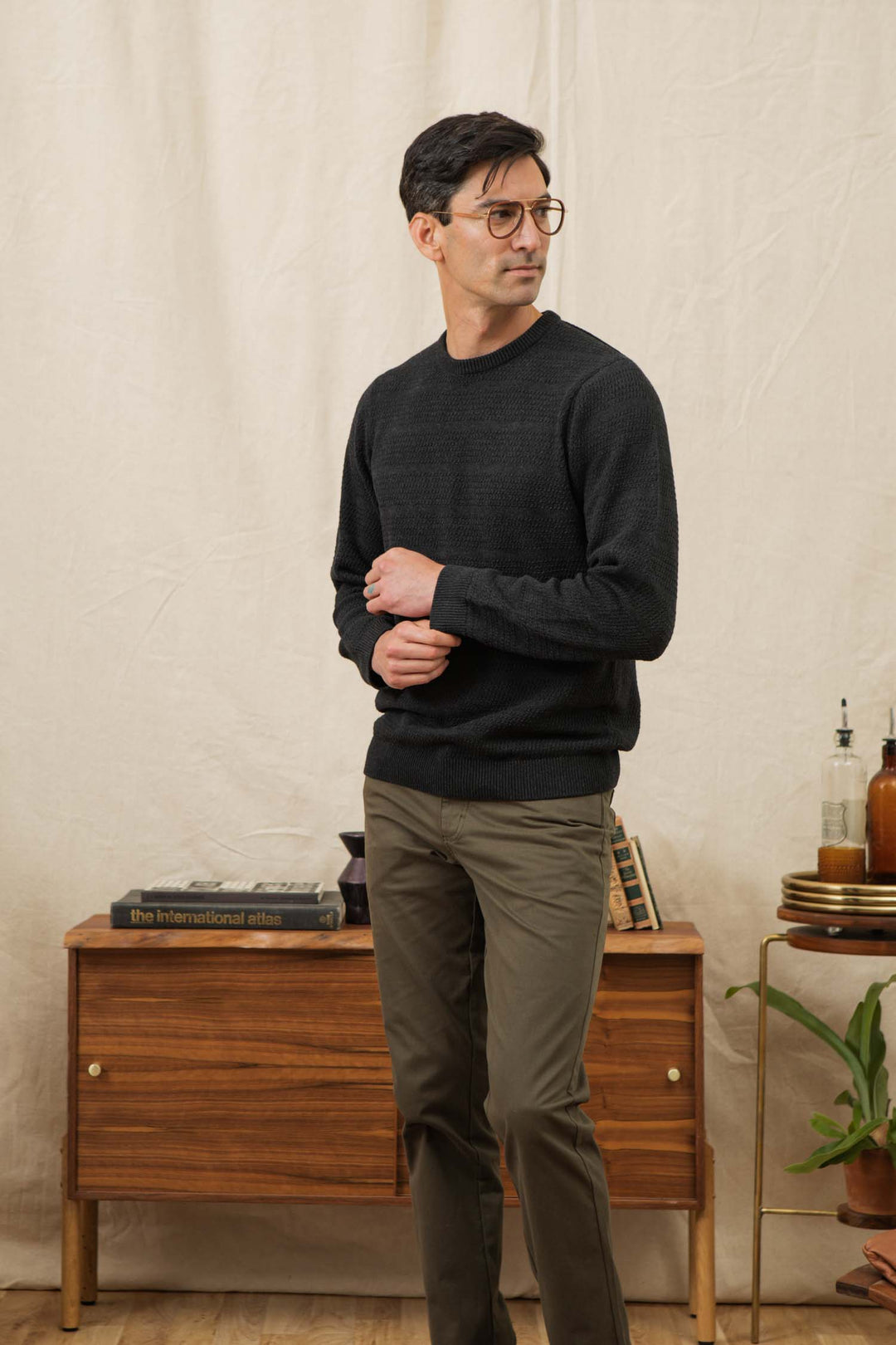 A man in glasses and olive pants stands indoors by a mid-century console, wearing the Bridge & Burn Calder Sweater in Midnight, looking to the side with a neutral expression.