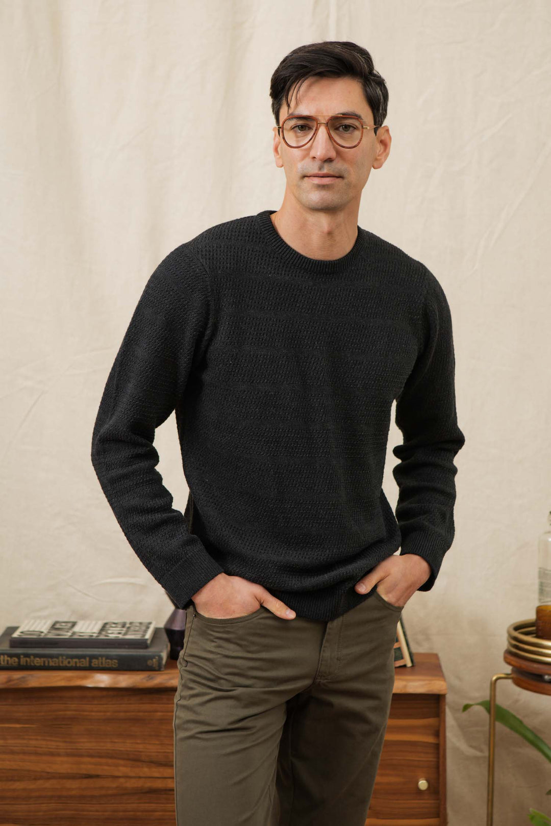 A man wearing glasses, the Bridge & Burn Calder Sweater in Midnight, and olive pants stands indoors with his hands in his pockets. Behind him is a wooden table with books and a glass bottle against a light fabric backdrop.