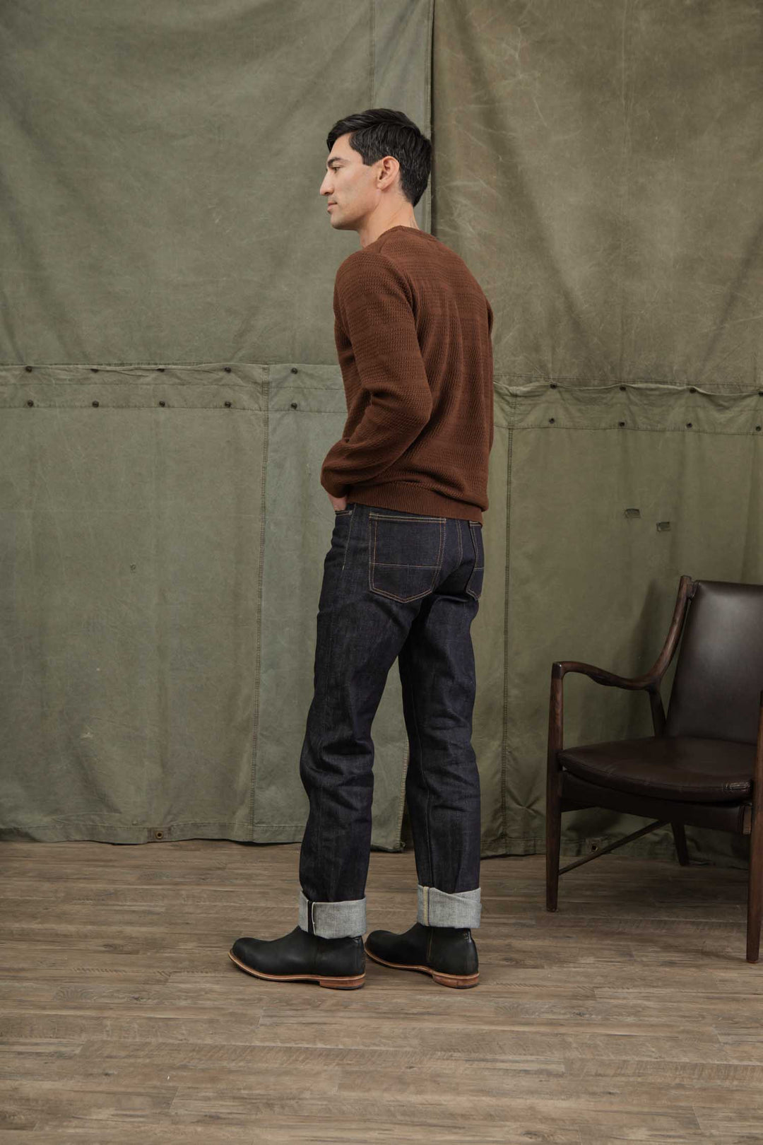 A man stands indoors in profile wearing the Bridge & Burn Calder Sweater in Walnut, paired with dark cuffed jeans and black boots. He faces left against a plain greenish backdrop, with a wooden chair to his right on a wooden floor.