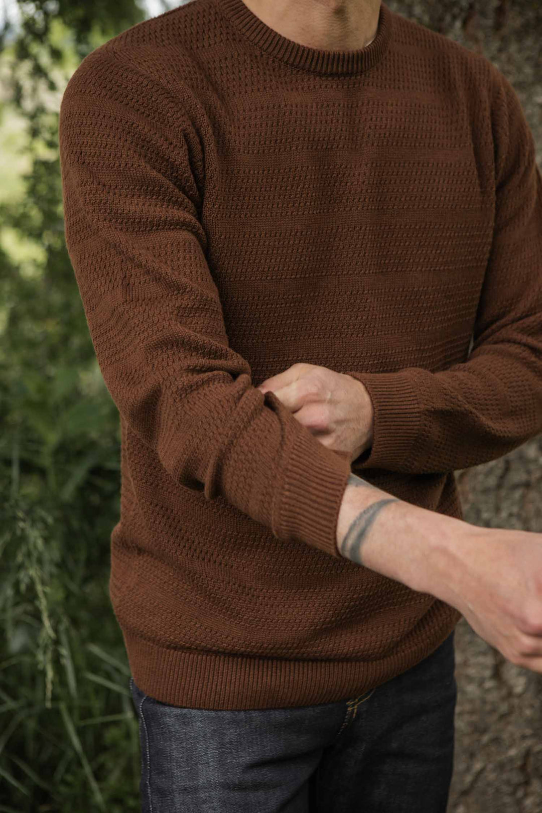 A person wearing the Bridge & Burn Calder Sweater in Walnut and jeans stands outdoors by greenery, rolling up their sleeve to reveal a forearm tattoo. Their face is not visible.