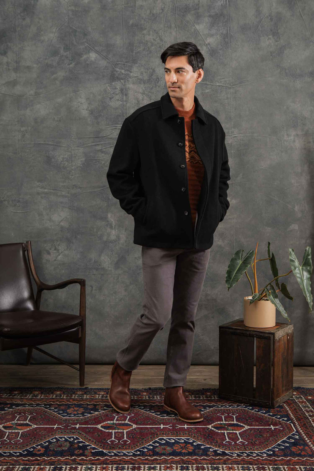 A man enjoys indoor comfort on a patterned rug, wearing the Bridge & Burn Clive Car Coat in black with his hands in his pockets, standing near a potted plant on a wooden box and a dark chair.