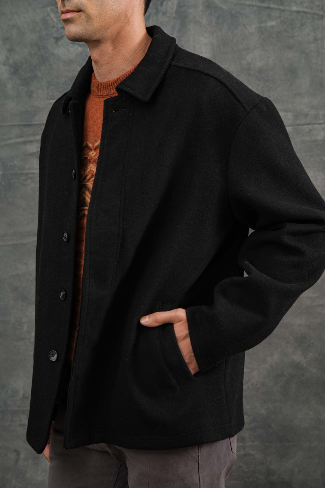 Wearing the Bridge & Burn Clive Car Coat in black over a brown sweater, a person stands with one hand in their pocket against a gray textured backdrop. Their face is partially out of frame, embodying effortless cold-weather style.