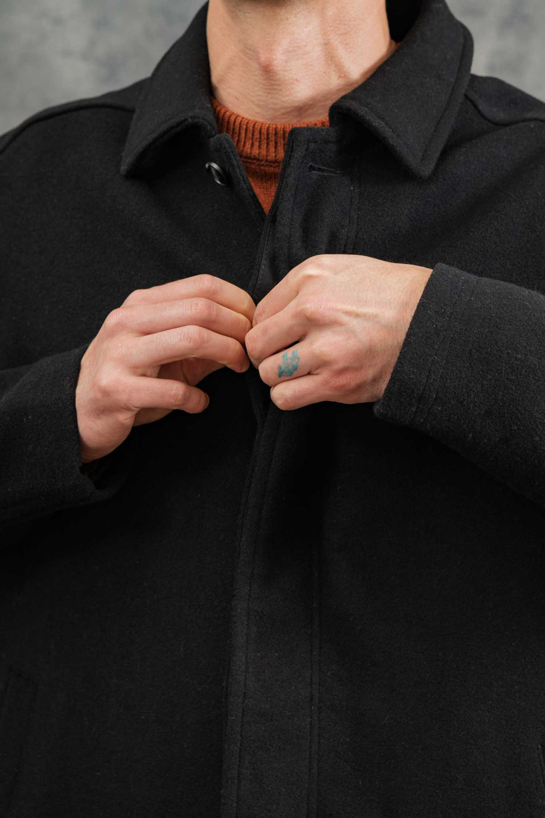 Wearing the Bridge & Burn Clive Car Coat in black over an orange sweater, a person buttons up for warmth; only their hands, chest, and lower face show, with a small blue tattoo on one finger.