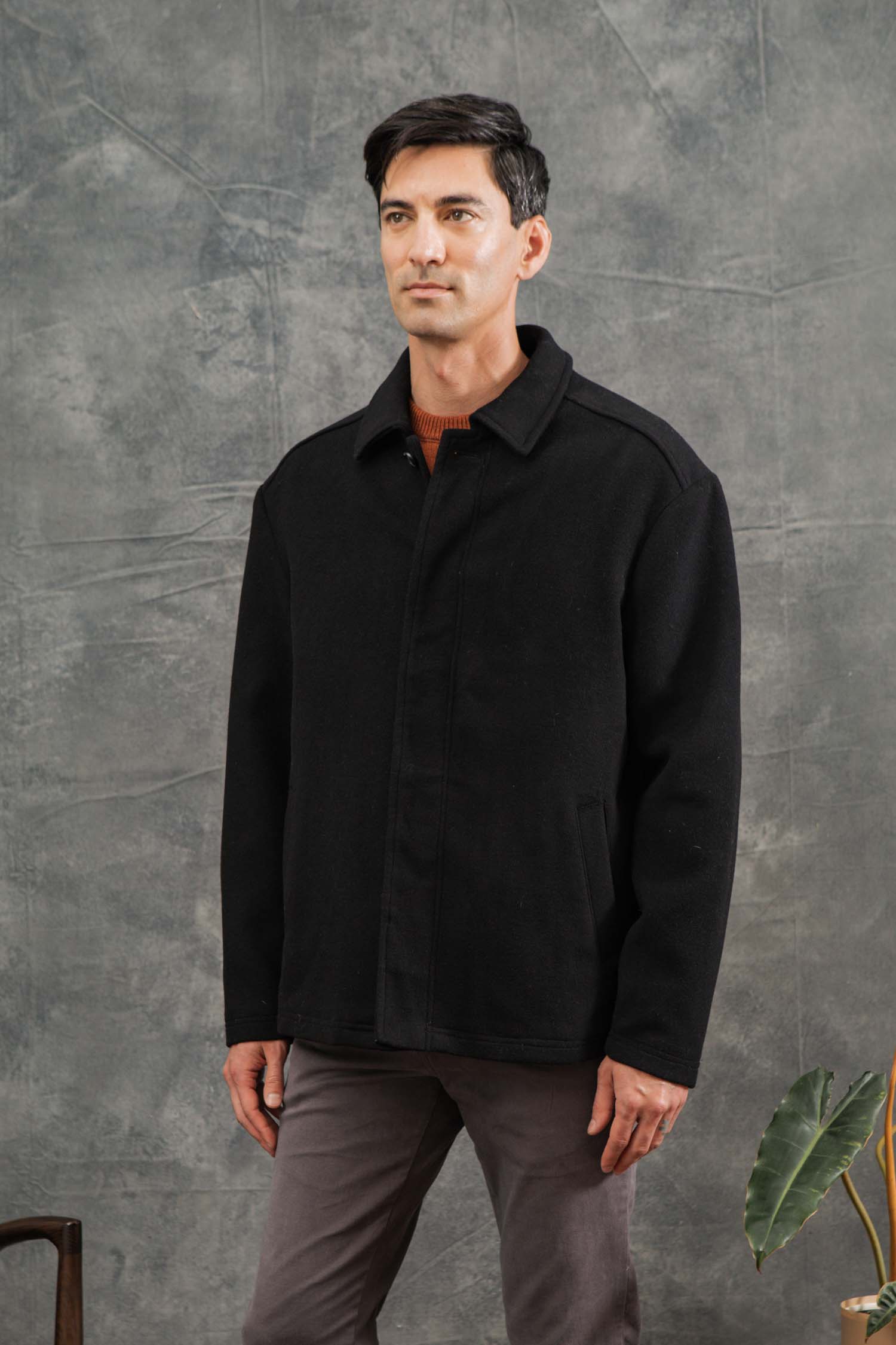 A man with short dark hair wears the Bridge & Burn Clive Car Coat in black over a brown top and gray pants, standing by a green potted plant in front of a textured gray backdrop.