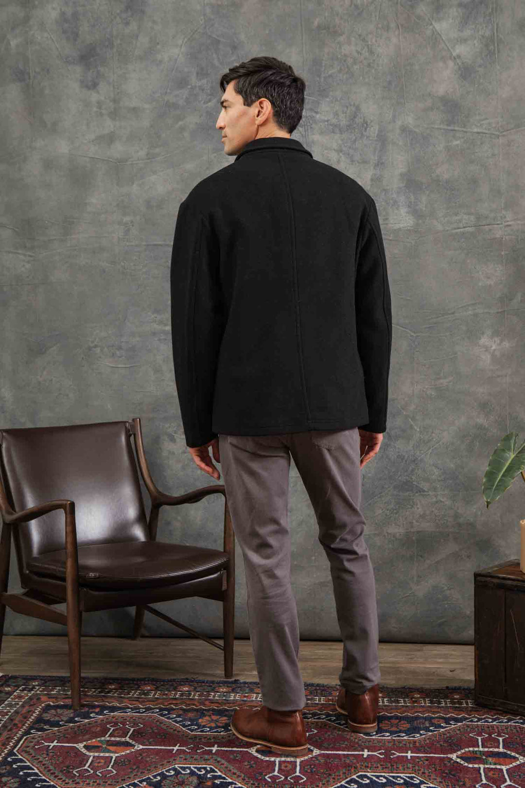 A man faces away from the camera in a Bridge & Burn Clive Car Coat in black, gray pants, and brown shoes. He stands beside a wooden chair with brown leather on a rug, set against a textured gray wall.