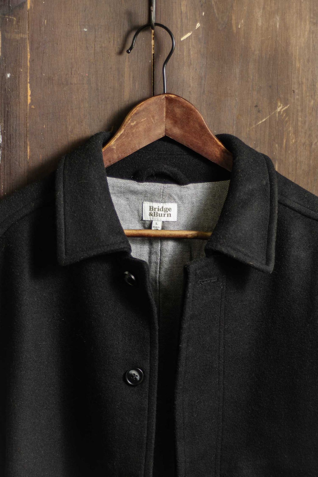 The Clive Car Coat in Black by Bridge & Burn hangs on a wooden hanger against a rustic wall, showcasing its heavyweight wool, collar design, black buttons, and visible label—perfect for timeless cold-weather style.