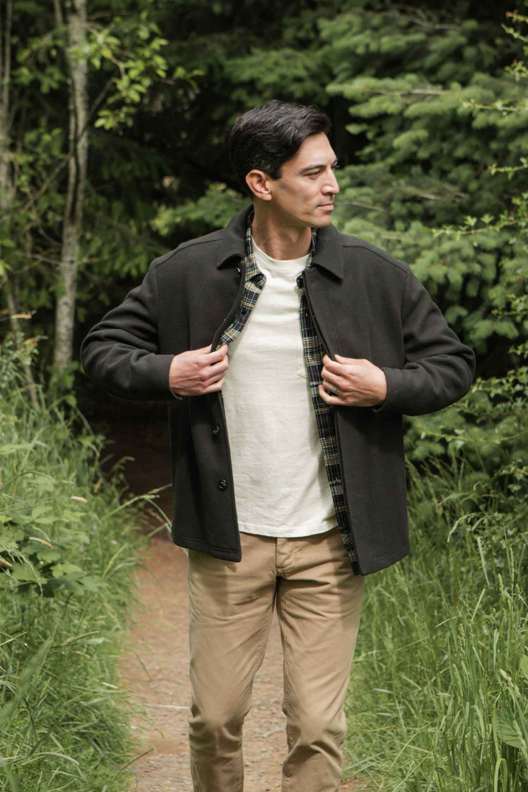 A man stands on a forest path, adjusting the buttons of his Clive Car Coat in Deep Sage by Bridge & Burn. He pairs it with a white shirt and tan pants, surrounded by lush green foliage and trees.