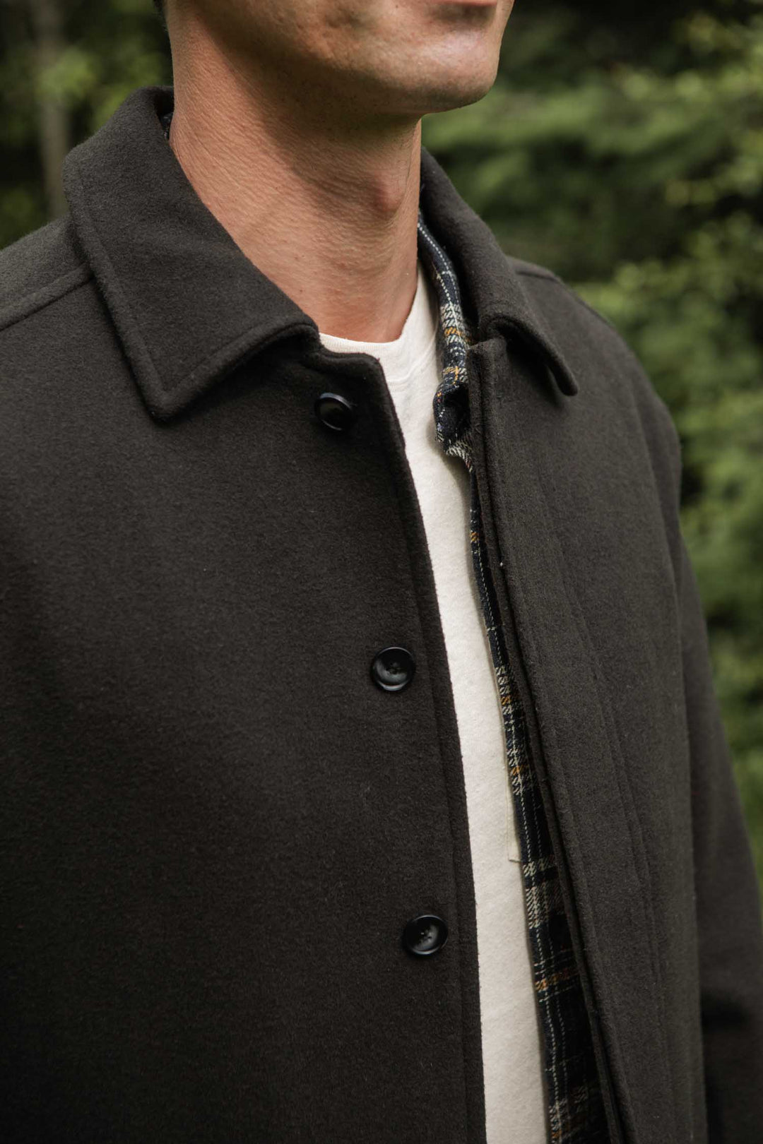Wearing the Bridge & Burn Clive Car Coat in Deep Sage over a plaid shirt and white tee, a person stands outdoors amid greenery, with only their lower face and upper torso visible.