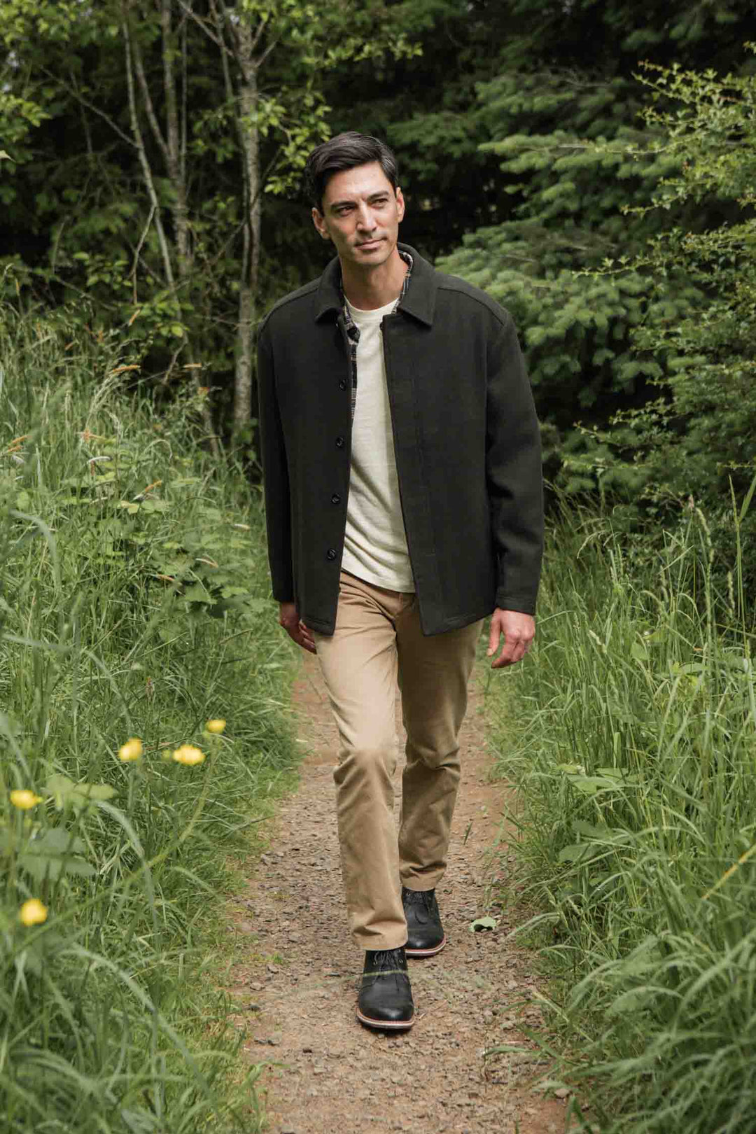 A man in the Bridge & Burn Clive Car Coat in Deep Sage, paired with a white shirt and khaki pants, strolls a narrow grassy forest trail lined with green trees and tall grass, enjoying cold-weather comfort.