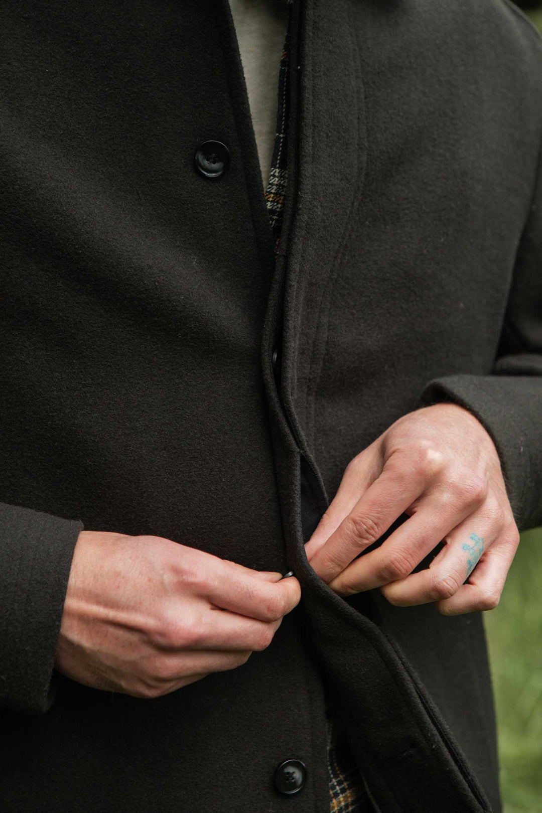Only hands and torso are visible as someone buttons the Bridge & Burn Clive Car Coat in Deep Sage, revealing a small blue finger tattoo. The blurred green backdrop adds a cozy, cold-weather vibe.
