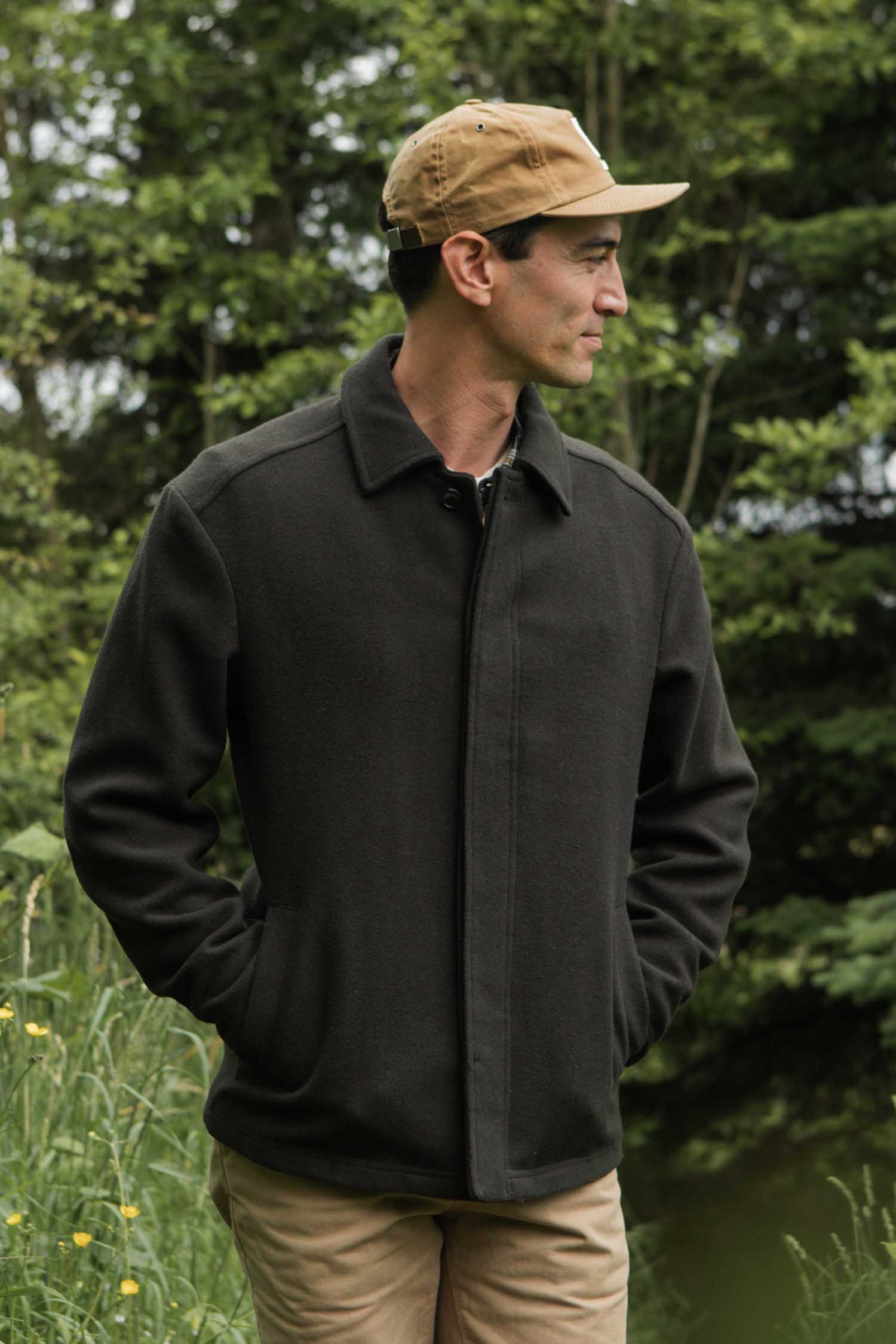 A man wearing the Bridge & Burn Clive Car Coat in Deep Sage stands outdoors in a grassy area with trees, looking to the side with his hands in his pockets.