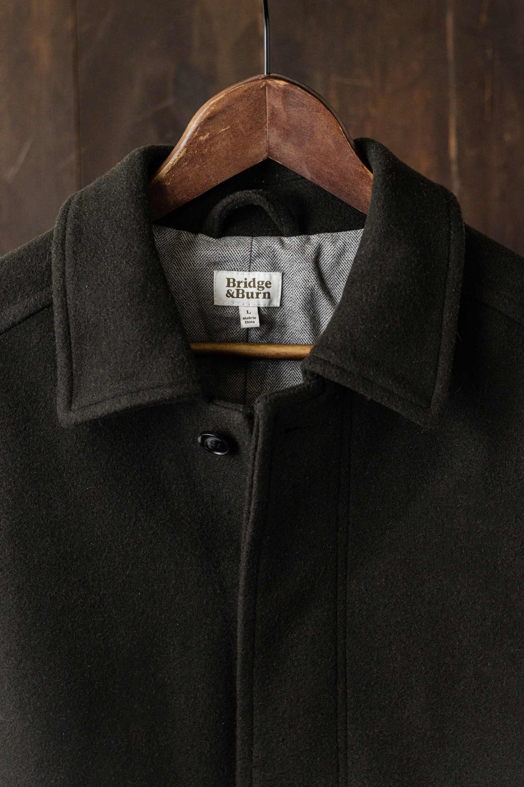 The Clive Car Coat in Deep Sage by Bridge & Burn hangs on a wooden hanger, buttoned at the top to reveal its gray lining and signature label. The dark wood backdrop highlights this heavyweight wool classic’s cozy, cold-weather appeal.