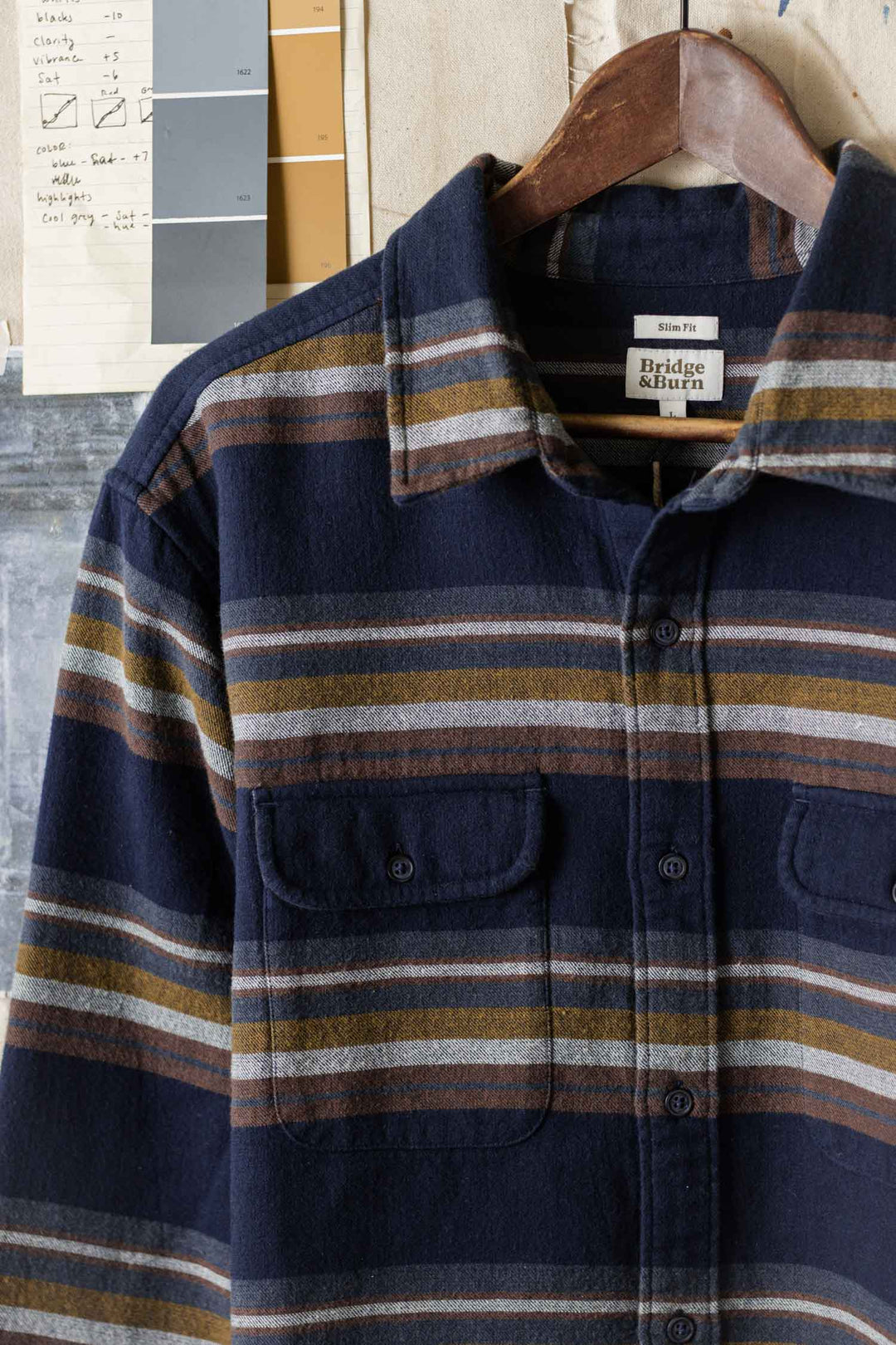 The Cole Slim Shirt / Coastline Stripe by Bridge & Burn—a navy, brown, and gray striped brushed cotton button-down with chest pockets—hangs on a wooden hanger against a wall featuring notepaper and a color swatch.