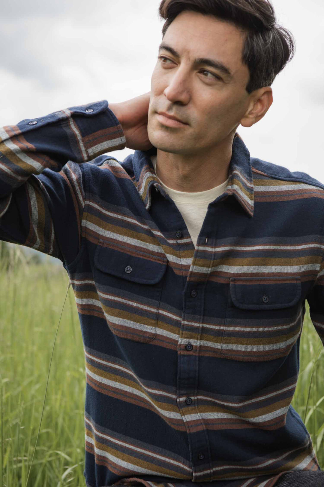 A man wearing the Bridge & Burn Cole Slim Shirt in Coastline Stripe sits in tall grass outdoors, one hand behind his neck as he gazes thoughtfully into the softly blurred, cloudy sky.