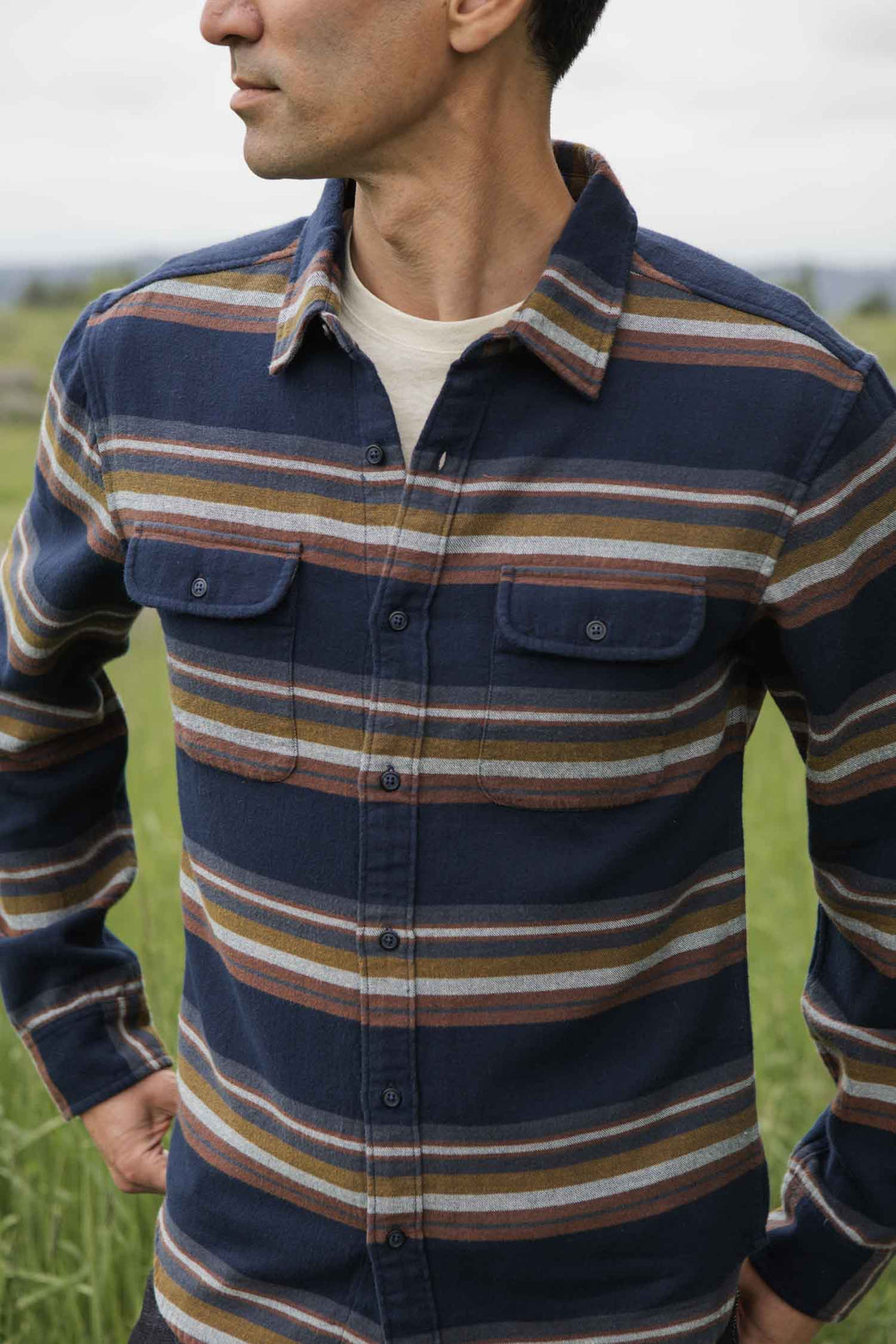 A man stands in a grassy field wearing the Bridge & Burn Cole Slim Shirt in Coastline Stripe, featuring a dark blue, slim fit design with horizontal brown, white, and gray stripes.