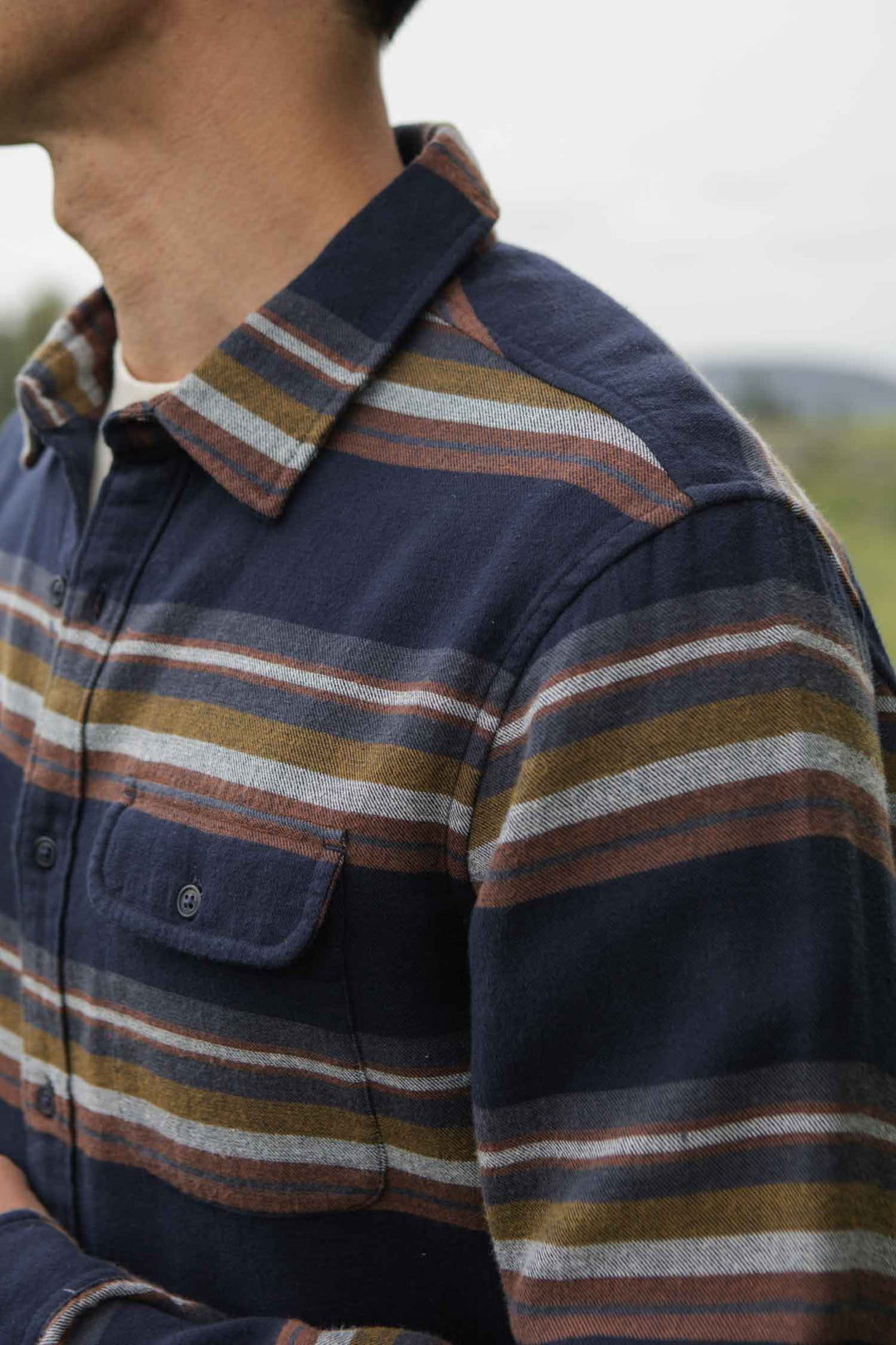 A person stands outdoors wearing the Bridge & Burn Cole Slim Shirt in Coastline Stripe—a navy brushed cotton flannel with brown, white, and olive stripes. The button-down collar and chest pocket are visible; the face is not shown.