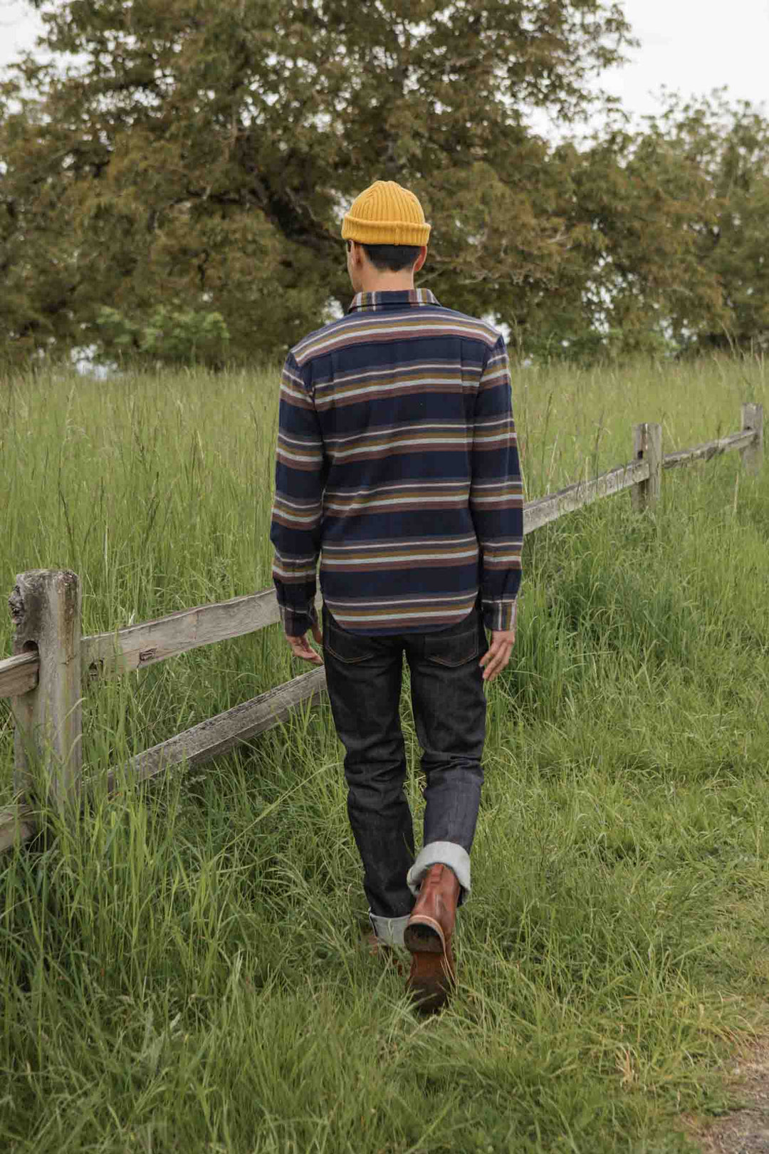 Wearing the Bridge & Burn Cole Slim Shirt in Coastline Stripe, a person in a yellow knit hat and rolled-up jeans walks on grass by a wooden fence, with tall green grass and a tree in the background.