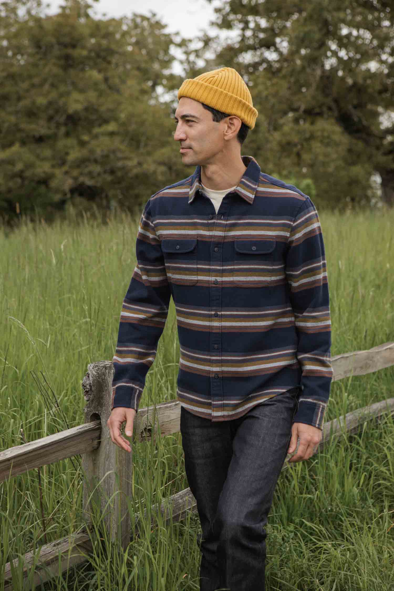 A person wearing the Bridge & Burn Cole Slim Shirt in Coastline Stripe walks outdoors by a wooden fence in a grassy field with trees in the background.