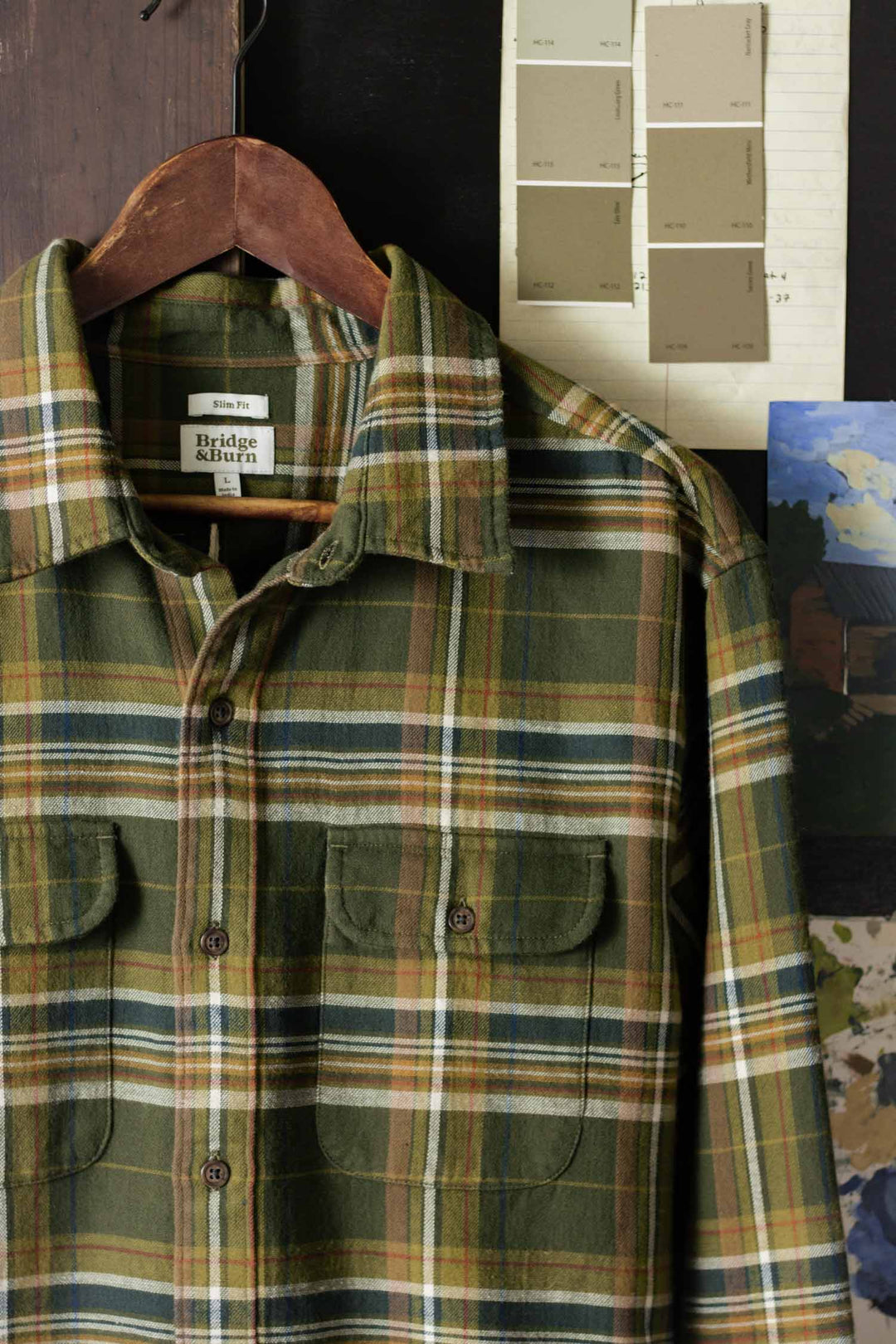 The Bridge & Burn Cole Slim Shirt in Green Mountain Plaid hangs on a wooden hanger, set against paint swatches, a rustic dark surface, and a partial painting of a building.