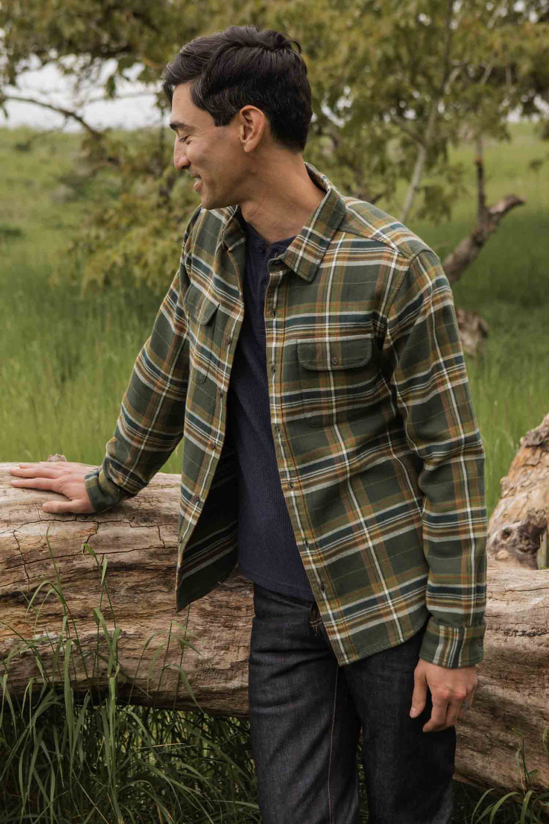 A man wearing the Bridge & Burn Cole Slim Shirt in Green Mountain Plaid stands outdoors, leaning on a fallen log and smiling with tall grass and a tree in the background.