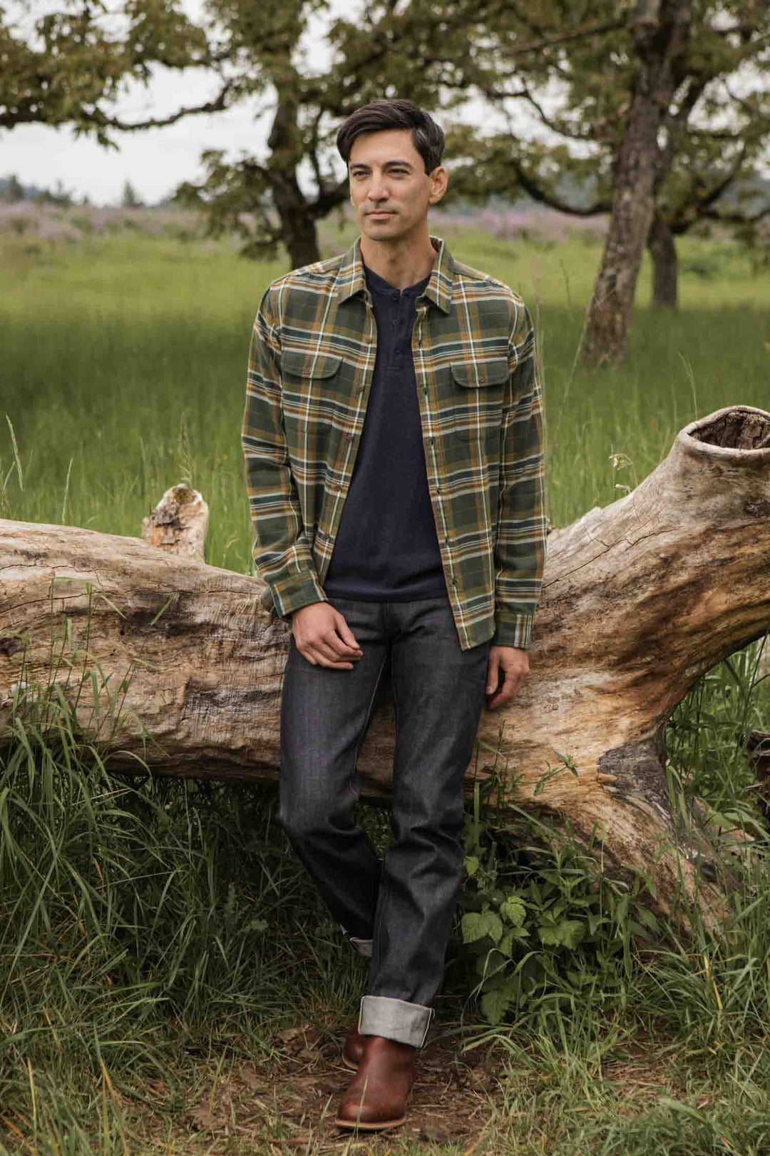 A man stands outdoors before a fallen tree, wearing Bridge & Burn's Cole Slim Shirt in Green Mountain Plaid, dark rolled-cuff jeans, and brown boots. The background has tall grass and leafy trees.