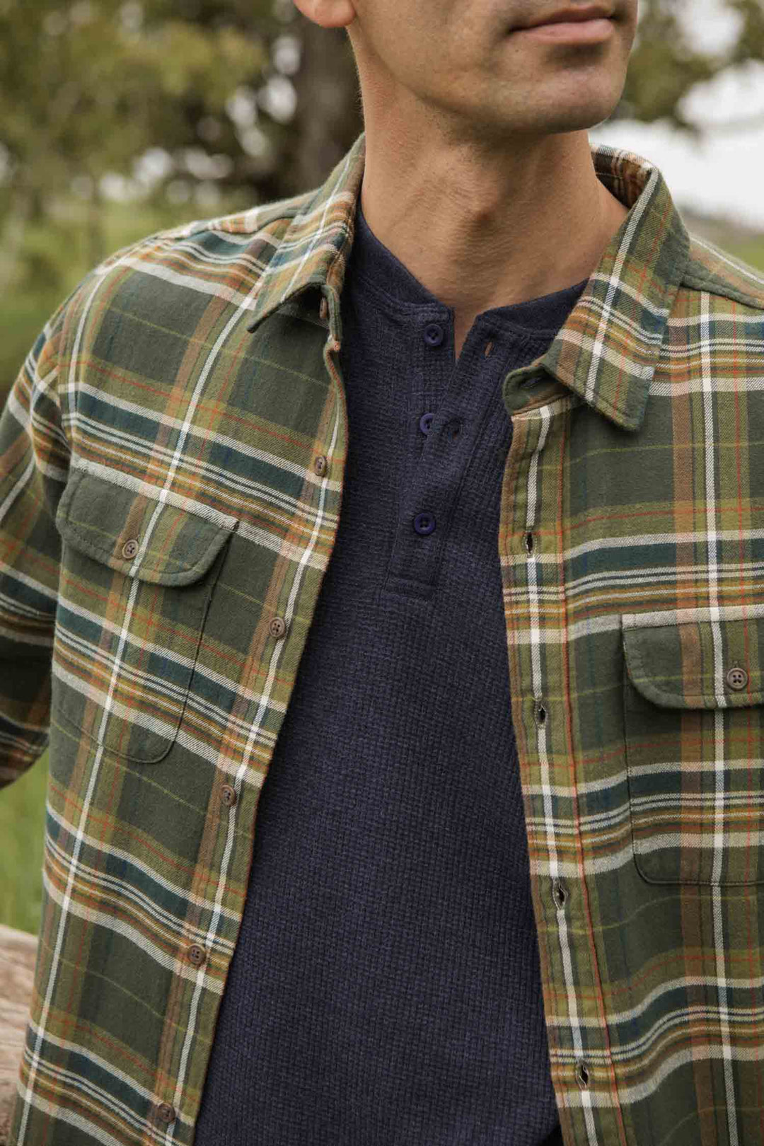 A person outdoors wears the Bridge & Burn Cole Slim Shirt in Green Mountain Plaid over a dark blue henley. The cropped image shows the torso and lower face with a blurred natural background.