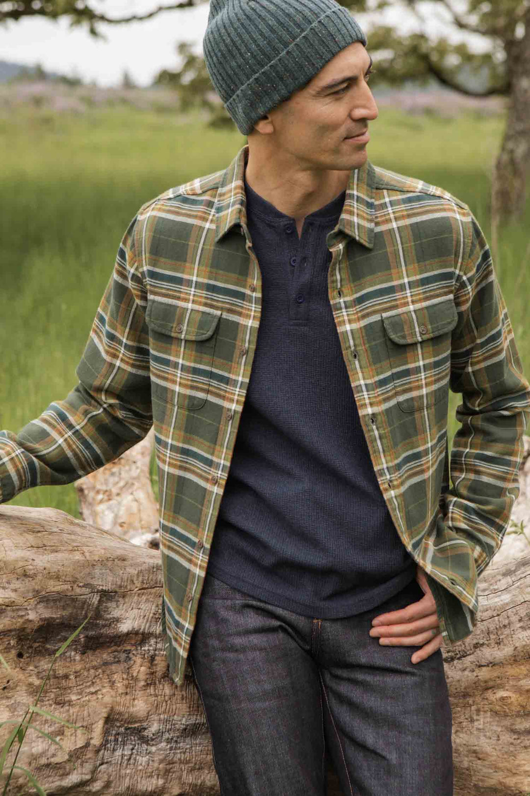 A man in the Bridge & Burn Cole Slim Shirt in Green Mountain Plaid stands outdoors by a fallen log, wearing a navy henley and green knit beanie, with grass and trees in the background.