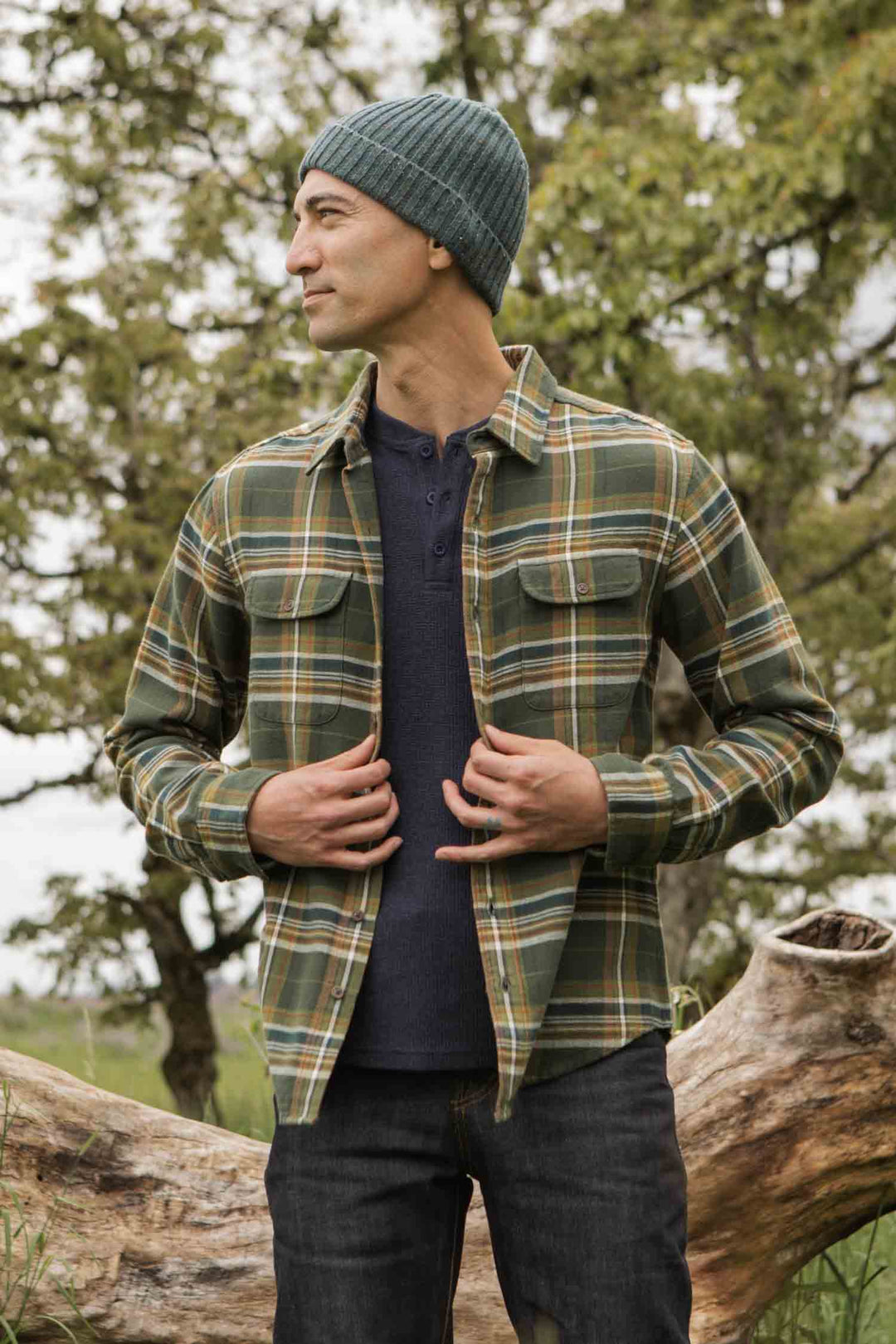 A man wearing the Bridge & Burn Cole Slim Shirt in Green Mountain Plaid, a blue knit beanie, and dark jeans stands outdoors by a fallen tree, looking to the side with a subtle smile. Lush trees and greenery form the background.