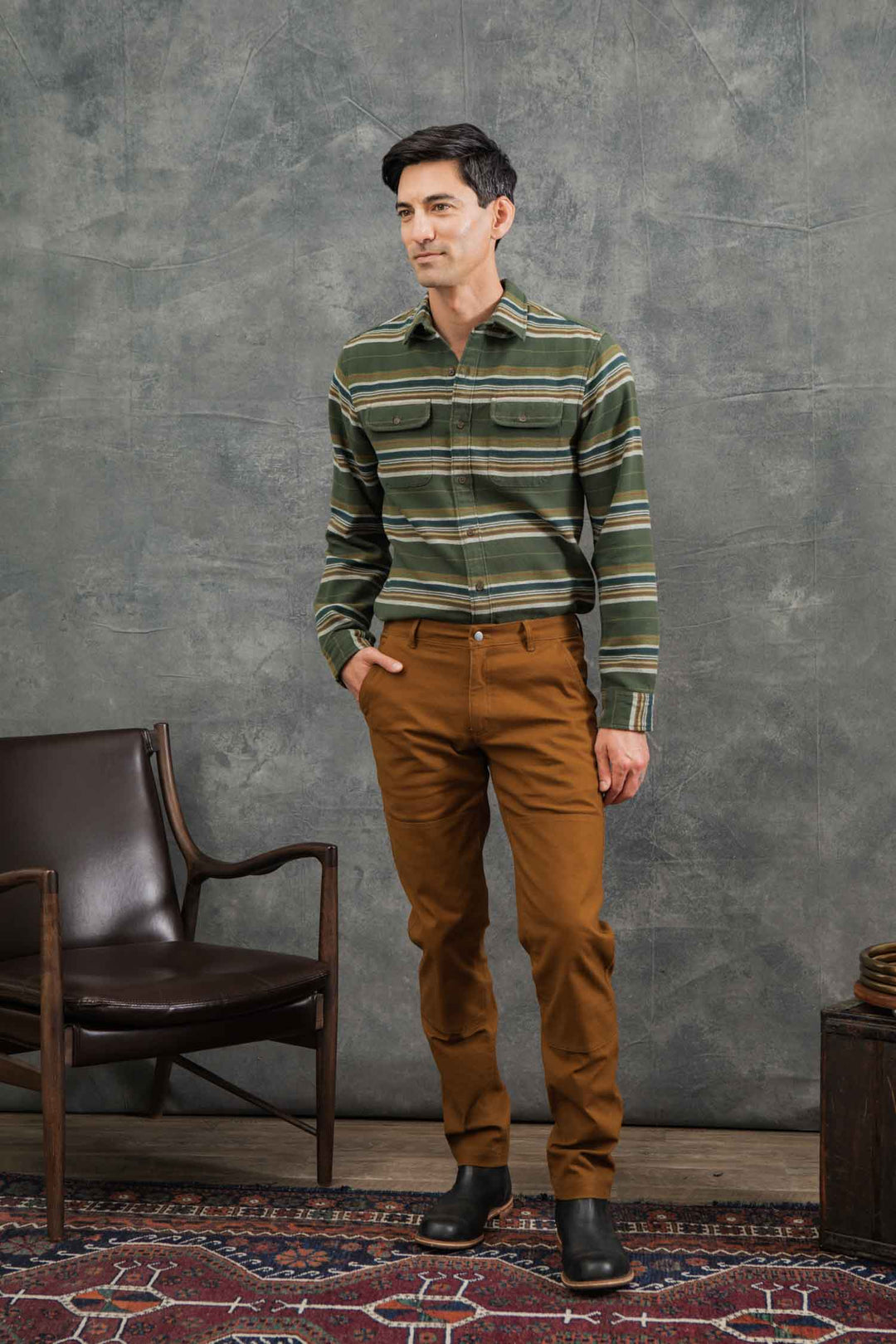 A man stands indoors on a patterned rug, wearing the Bridge & Burn Cole Slim Shirt in Wreath Stripe, brown pants, and black boots with one hand in his pocket. A brown armchair and small table are beside him.