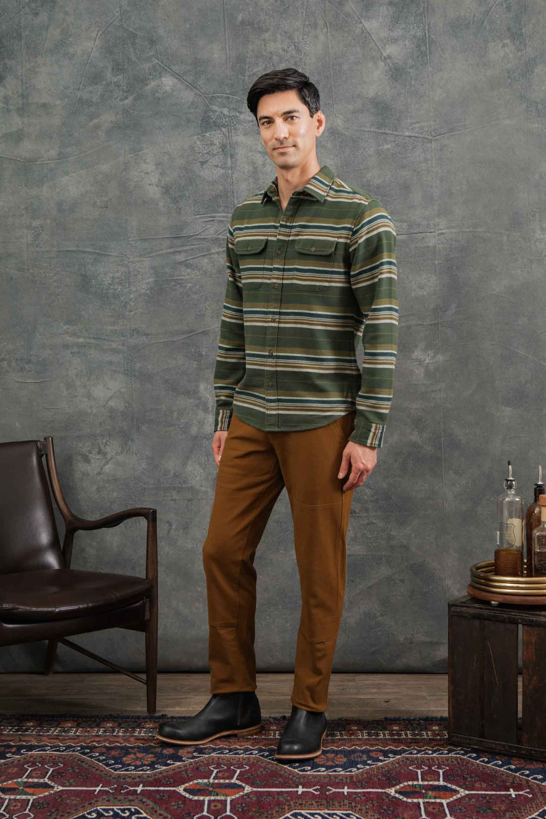 A man stands indoors on a patterned rug, wearing Bridge & Burn's Cole Slim Shirt in Wreath Stripe and brown pants, with black boots. He is beside a dark chair and small table with bottles, set against a textured gray backdrop.