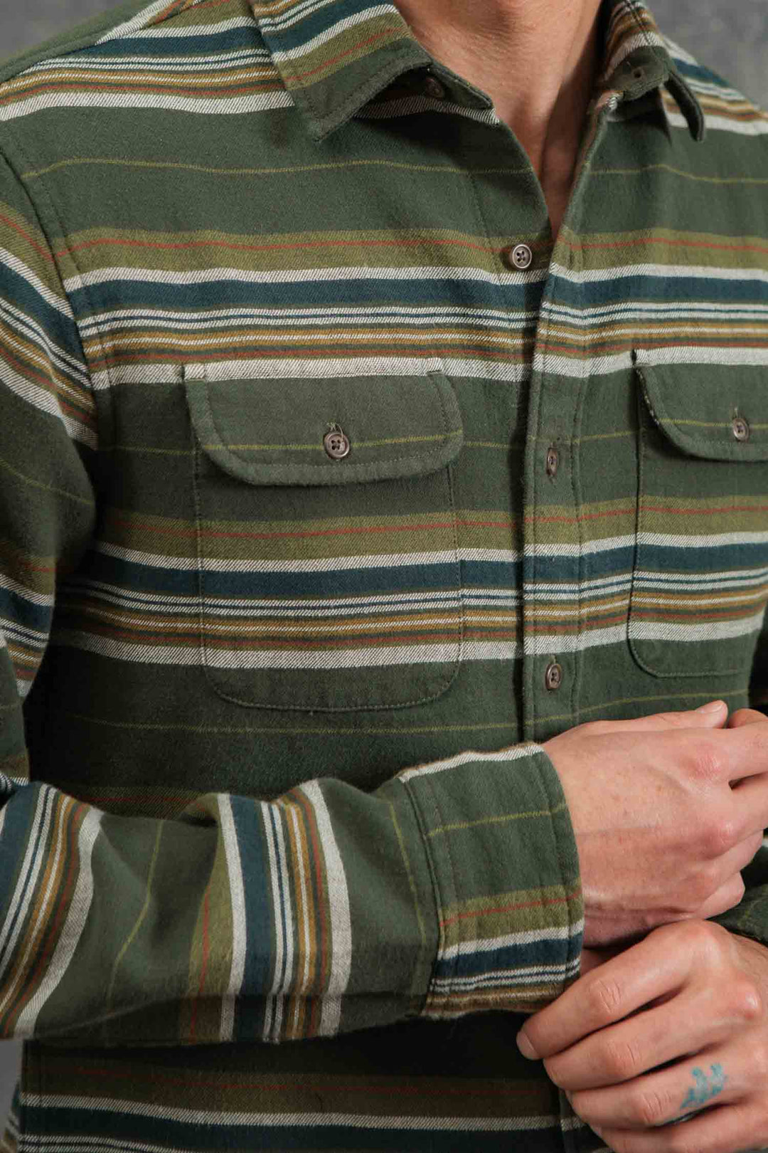 A person wears the Bridge & Burn Cole Slim Shirt in Wreath Stripe, a green, button-down flannel with blue, yellow, and white horizontal stripes made of soft brushed cotton. Their hands are visible, one resting on the other.