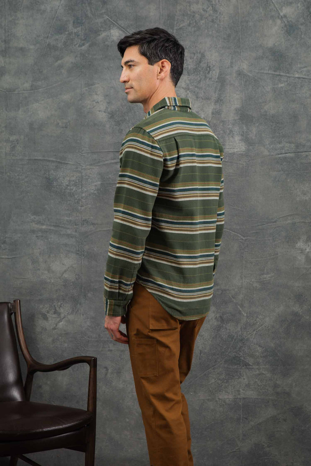 A man with short dark hair stands sideways, facing left. He wears the Bridge & Burn Cole Slim Shirt in Wreath Stripe and brown pants. A dark chair is to his left against a textured gray background.