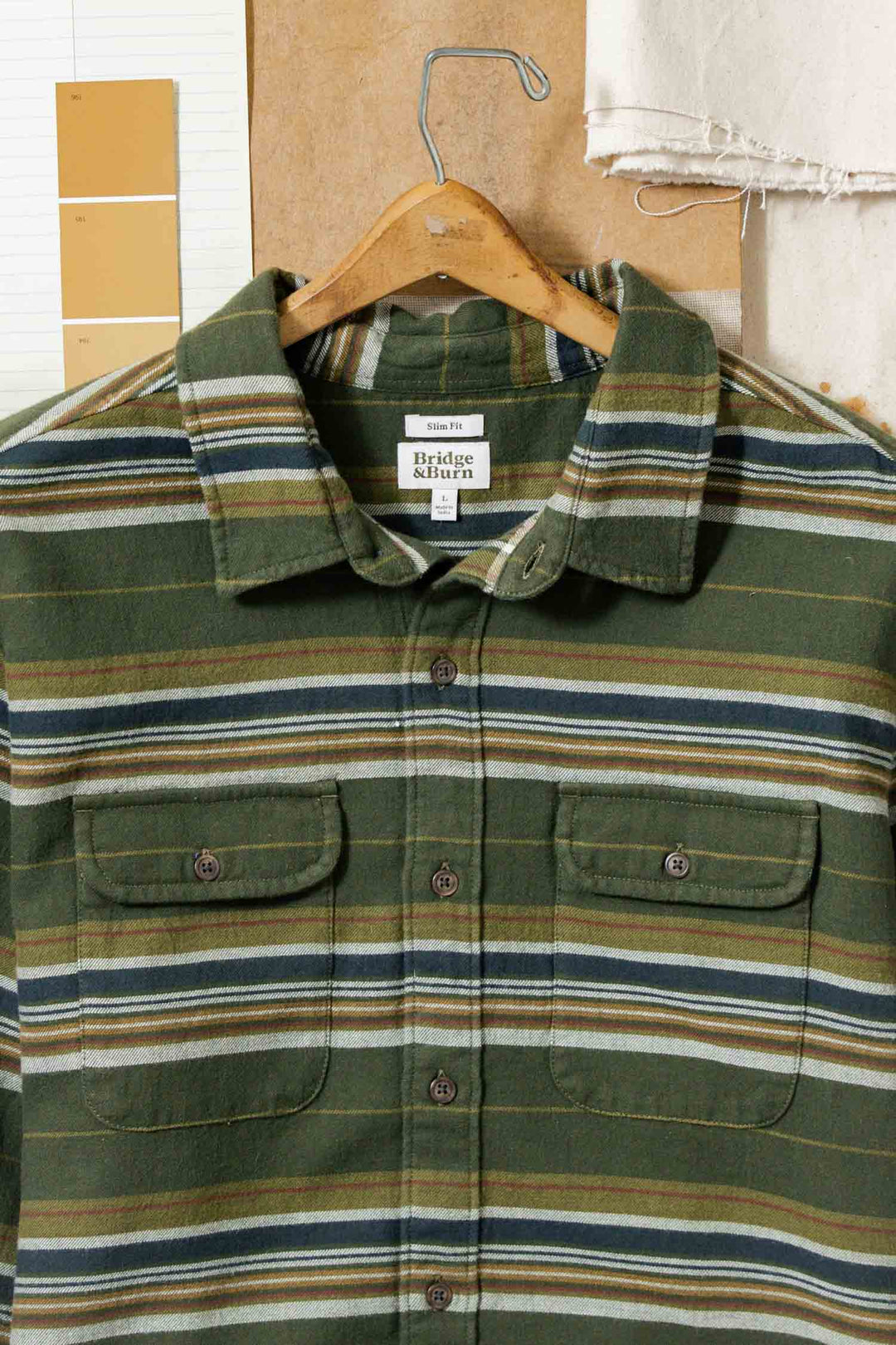 The Cole Slim Shirt / Wreath Stripe by Bridge & Burn, featuring green, blue, white, and brown stripes in brushed cotton, hangs on a wooden hanger against a brown background with fabric swatches and paper samples behind it.
