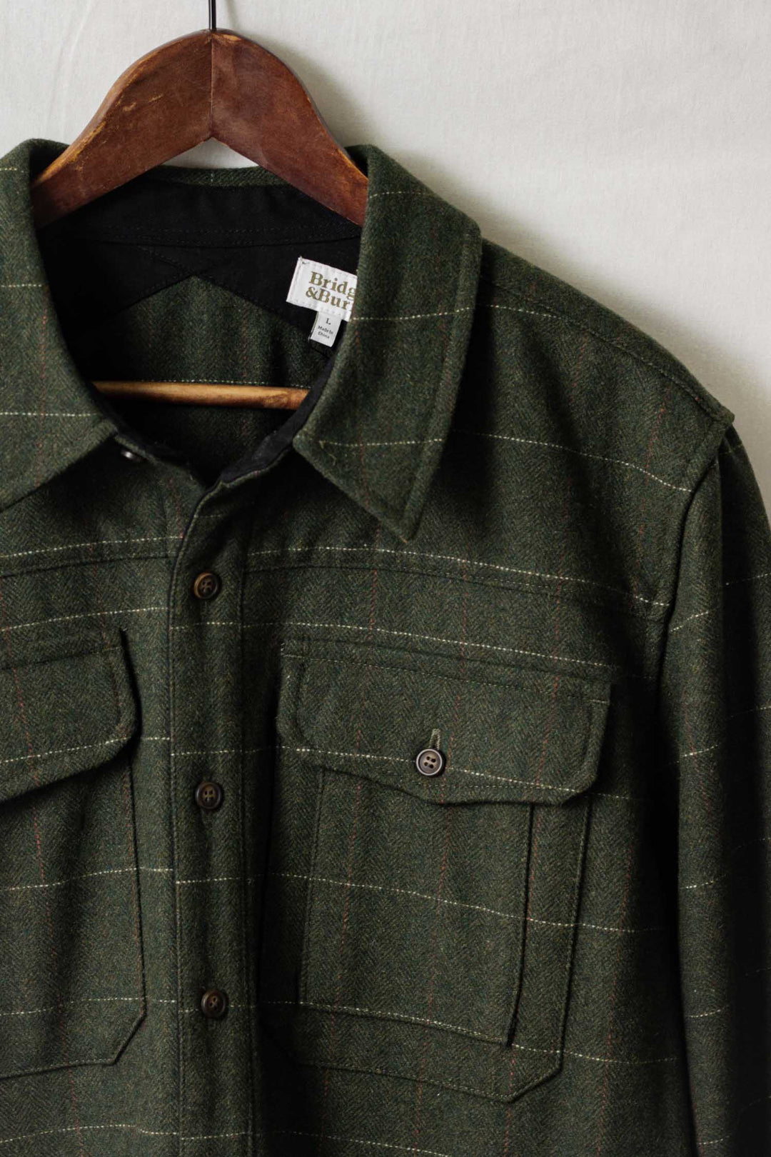 The Bridge & Burn Fielding Shirt Jacket in Cypress Windowpane hangs on a wooden hanger against a light background, featuring flap chest pockets, dark buttons, and a classic green plaid check for timeless overshirt style.