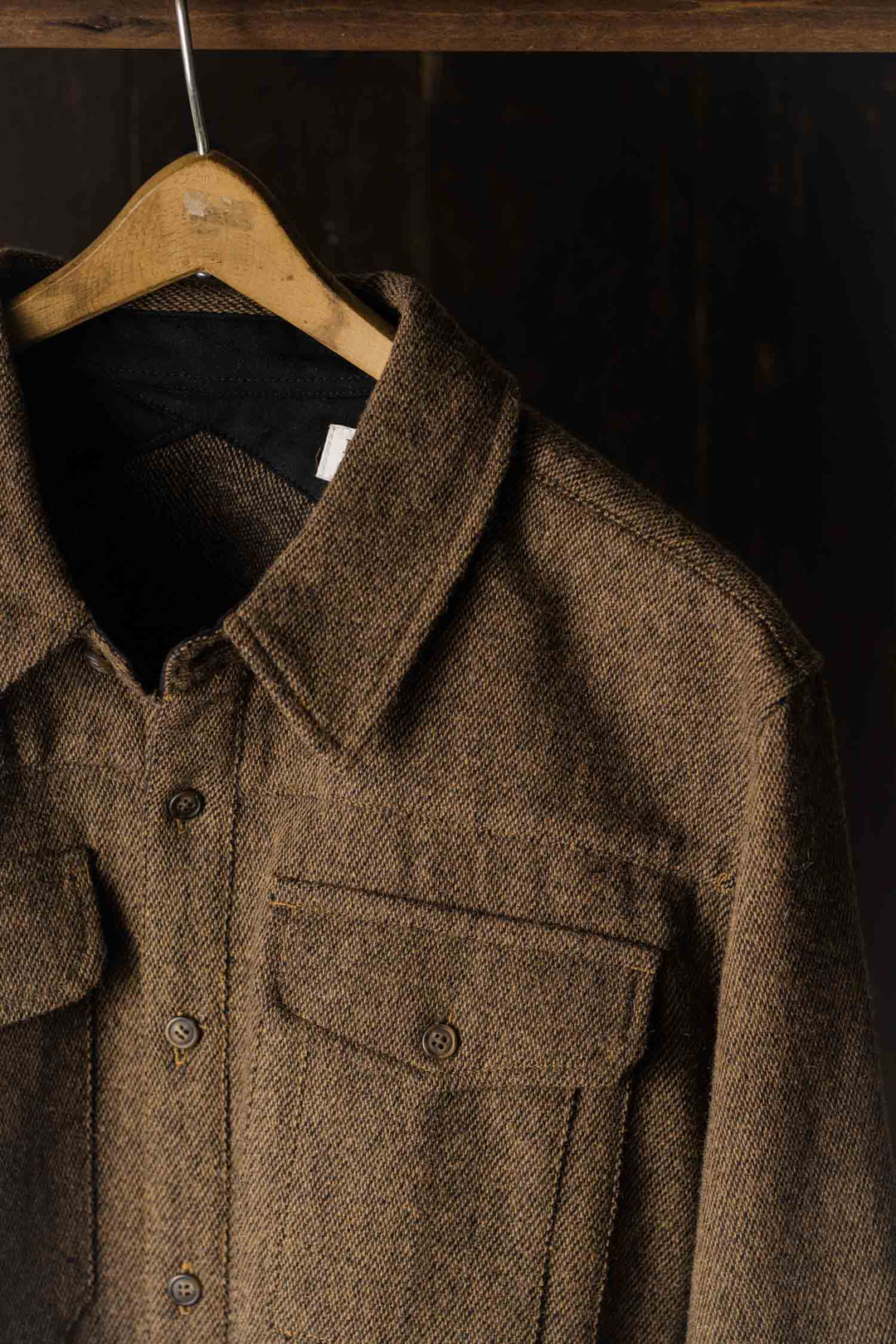 A close-up of the Bridge & Burn Fielding Shirt Jacket in Tawny Tweed on a wooden hanger, highlighting its collar, buttons, and front chest pockets—this classic overshirt pops against a dark background.