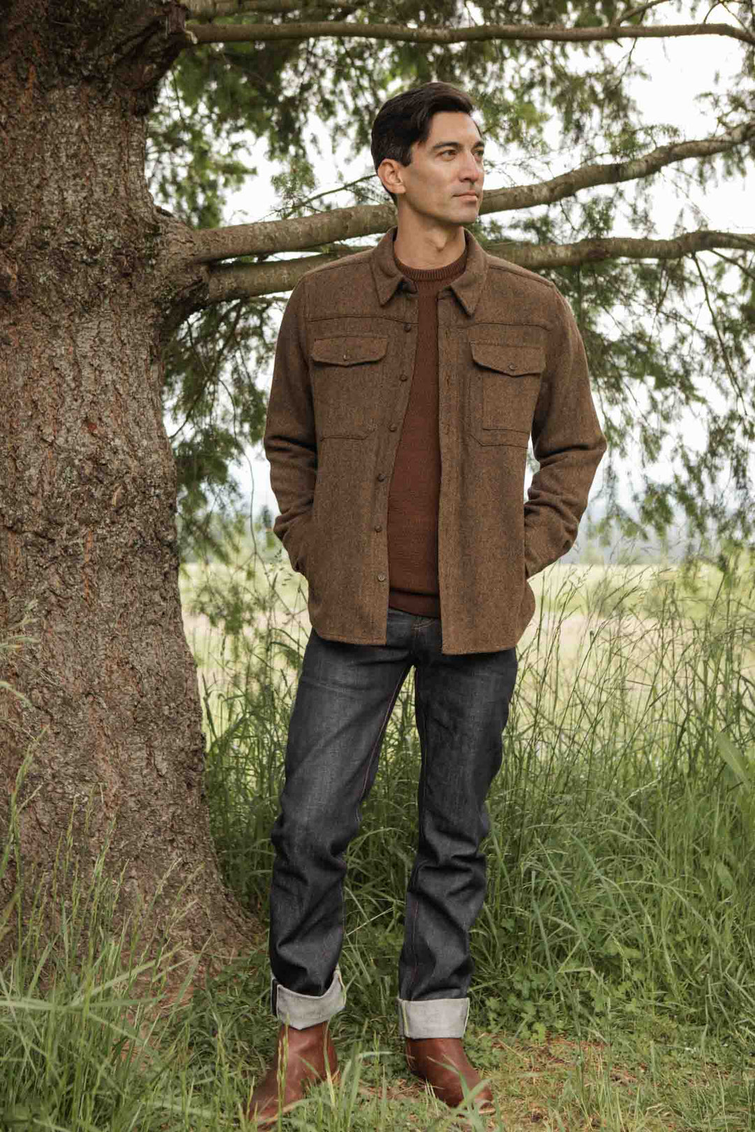 A man stands in tall grass by a tree, hands in his pockets, wearing the Bridge & Burn Fielding Shirt Jacket in Tawny Tweed over dark jeans and brown boots, glancing to the side.