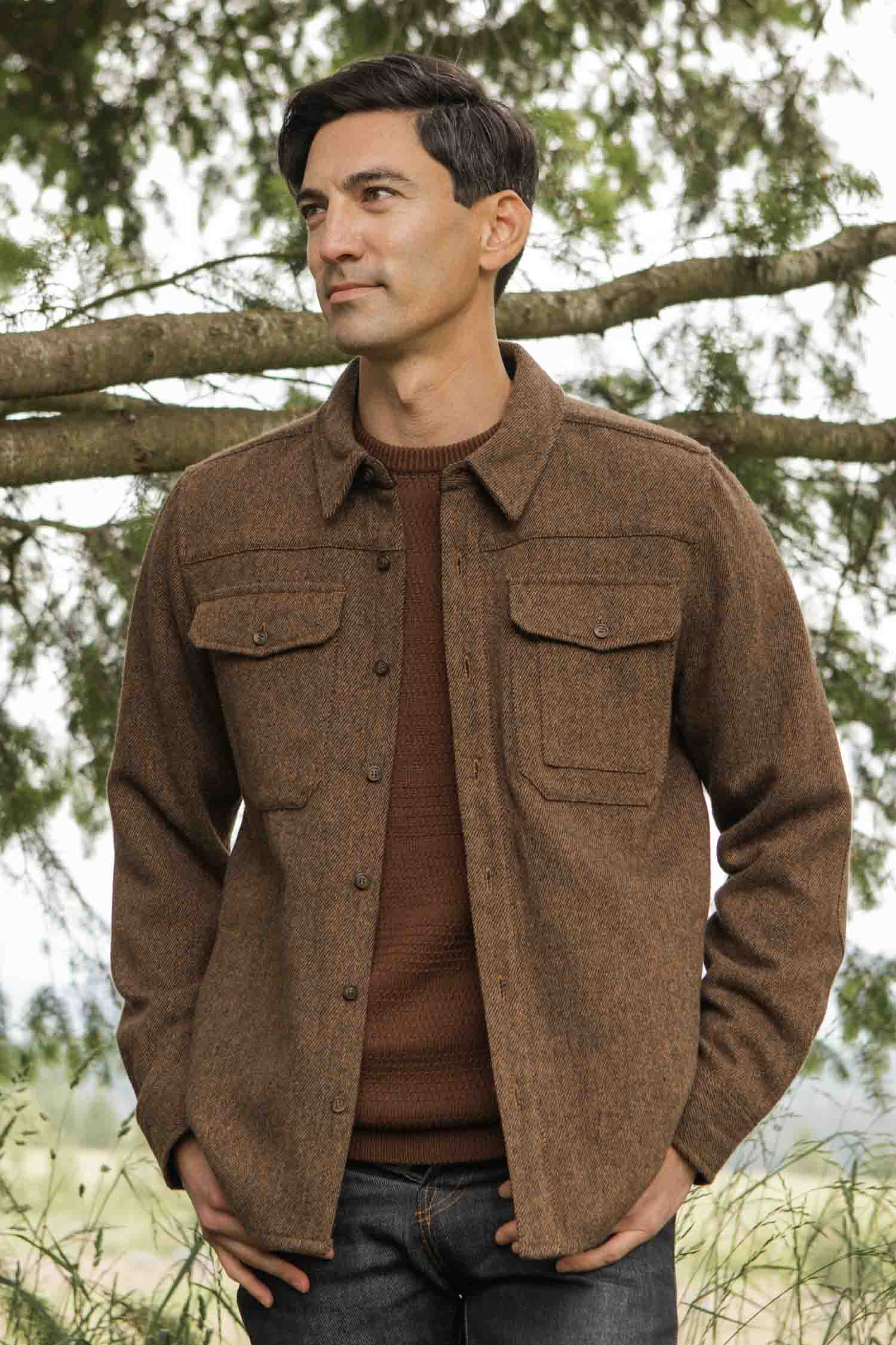 A man stands outdoors before pine branches, wearing the Bridge & Burn Fielding Shirt Jacket in Tawny Tweed over a brown sweater and dark jeans. He gazes off to the side with a slight smile.