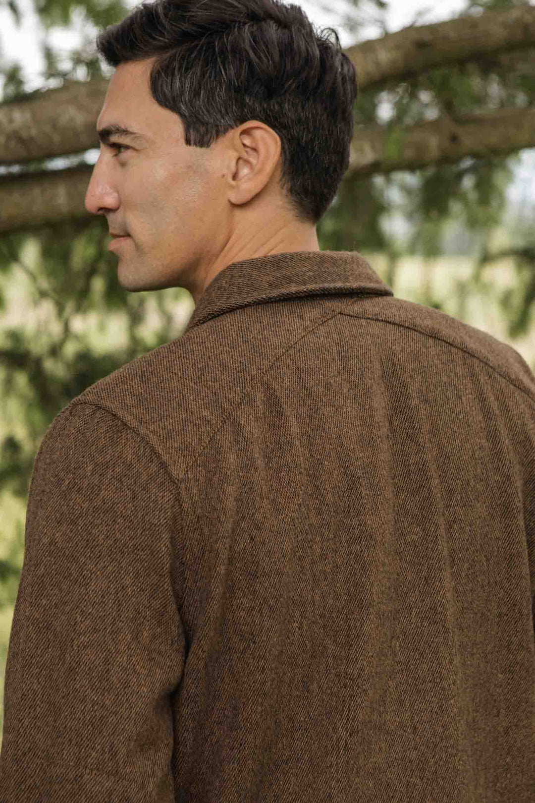 A man with short dark hair stands outdoors, looking to the side. He wears the Bridge & Burn Fielding Shirt Jacket in Tawny Tweed, surrounded by green trees and blurred natural scenery in the background.