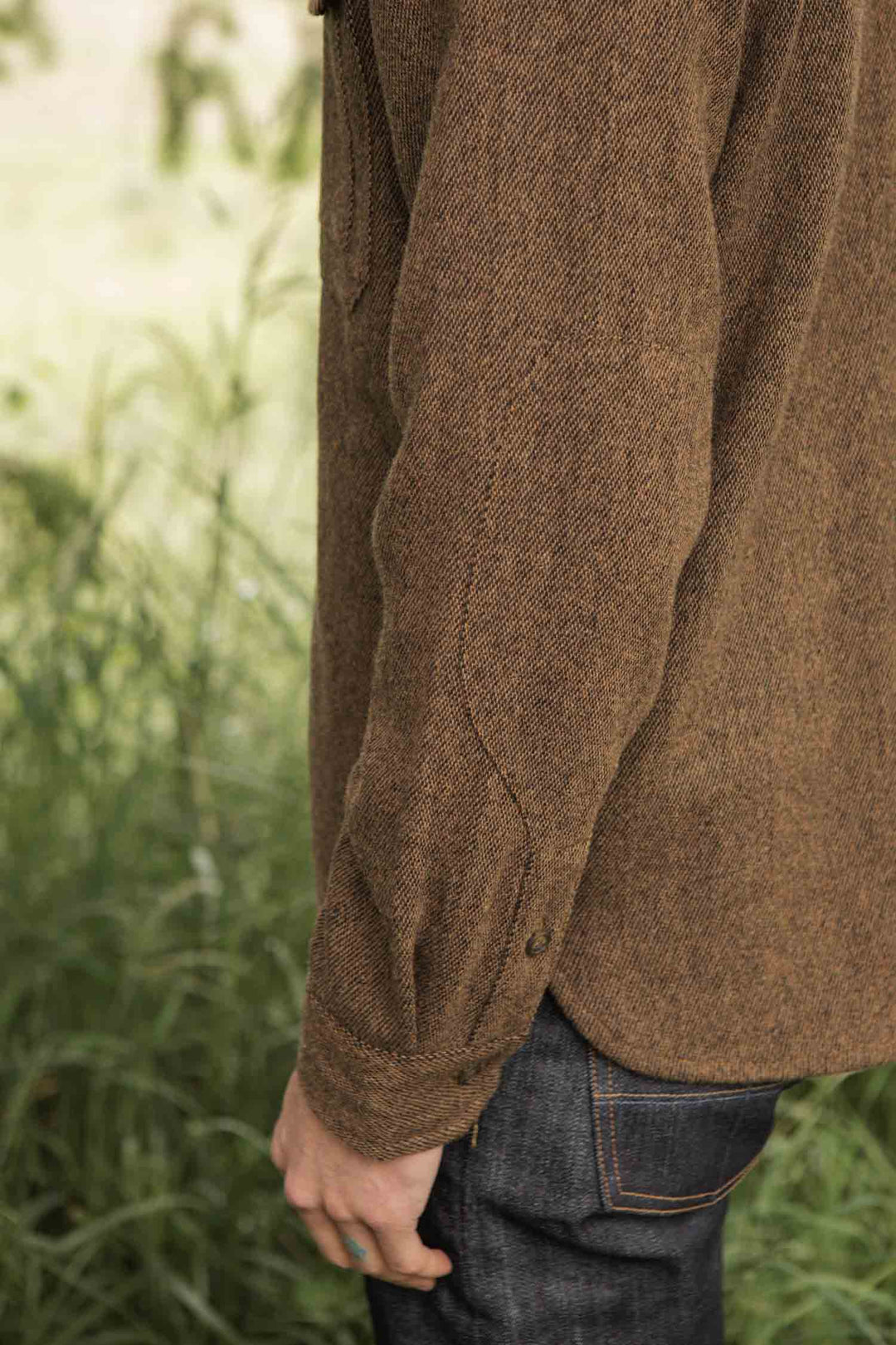 A person stands in tall grass wearing the Bridge & Burn Fielding Shirt Jacket in Tawny Tweed and dark denim jeans, with only their arm and side visible.