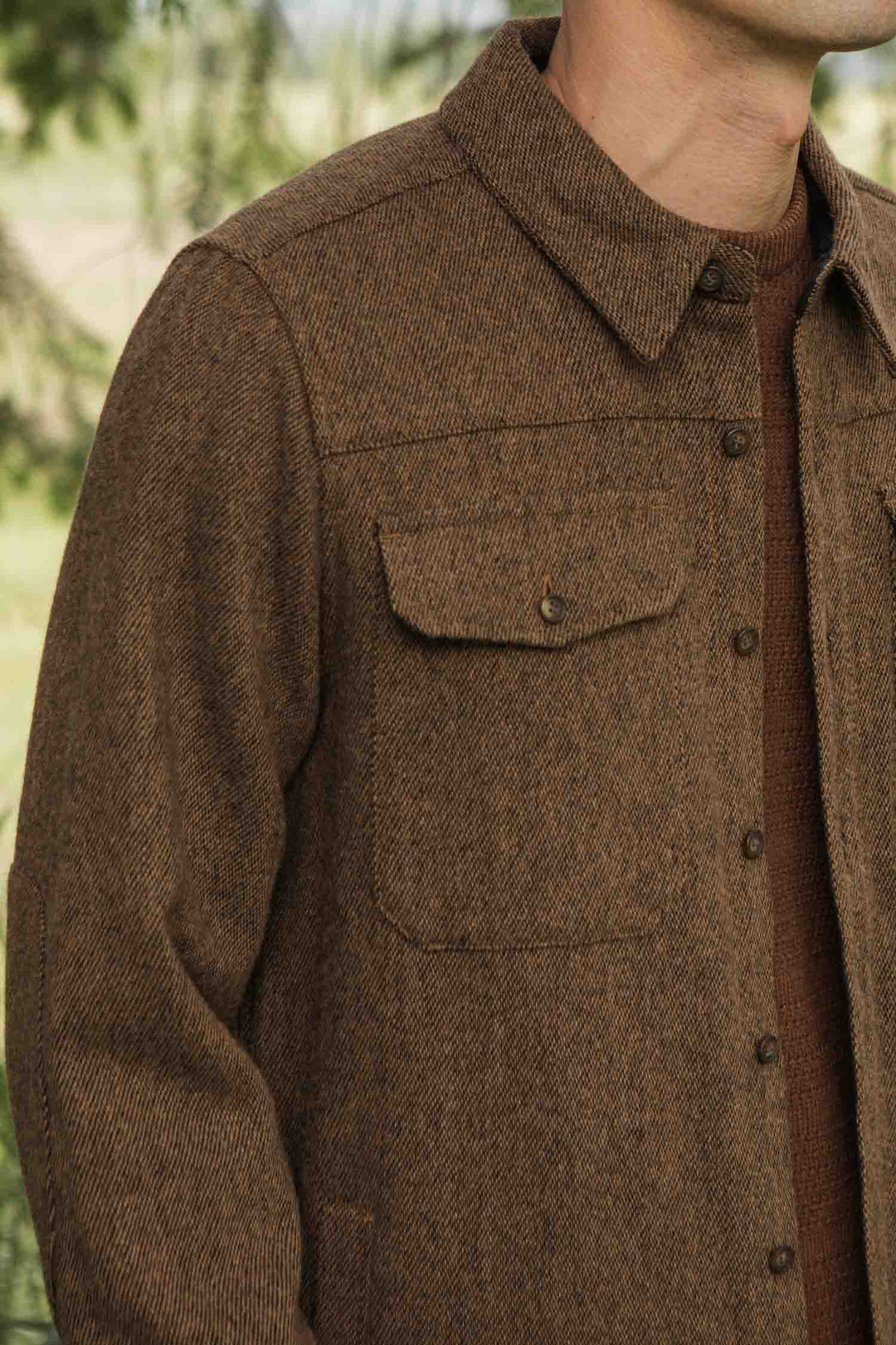 A person wears the Bridge & Burn Fielding Shirt Jacket in Tawny Tweed, featuring front chest pockets, while standing outdoors. Only their upper torso and part of their face are visible.