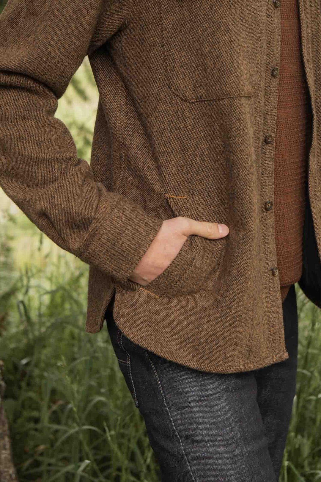 A person stands outdoors with one hand in the pocket of a Bridge & Burn Fielding Shirt Jacket in Tawny Tweed, paired with dark jeans. Grass and greenery can be seen in the background.