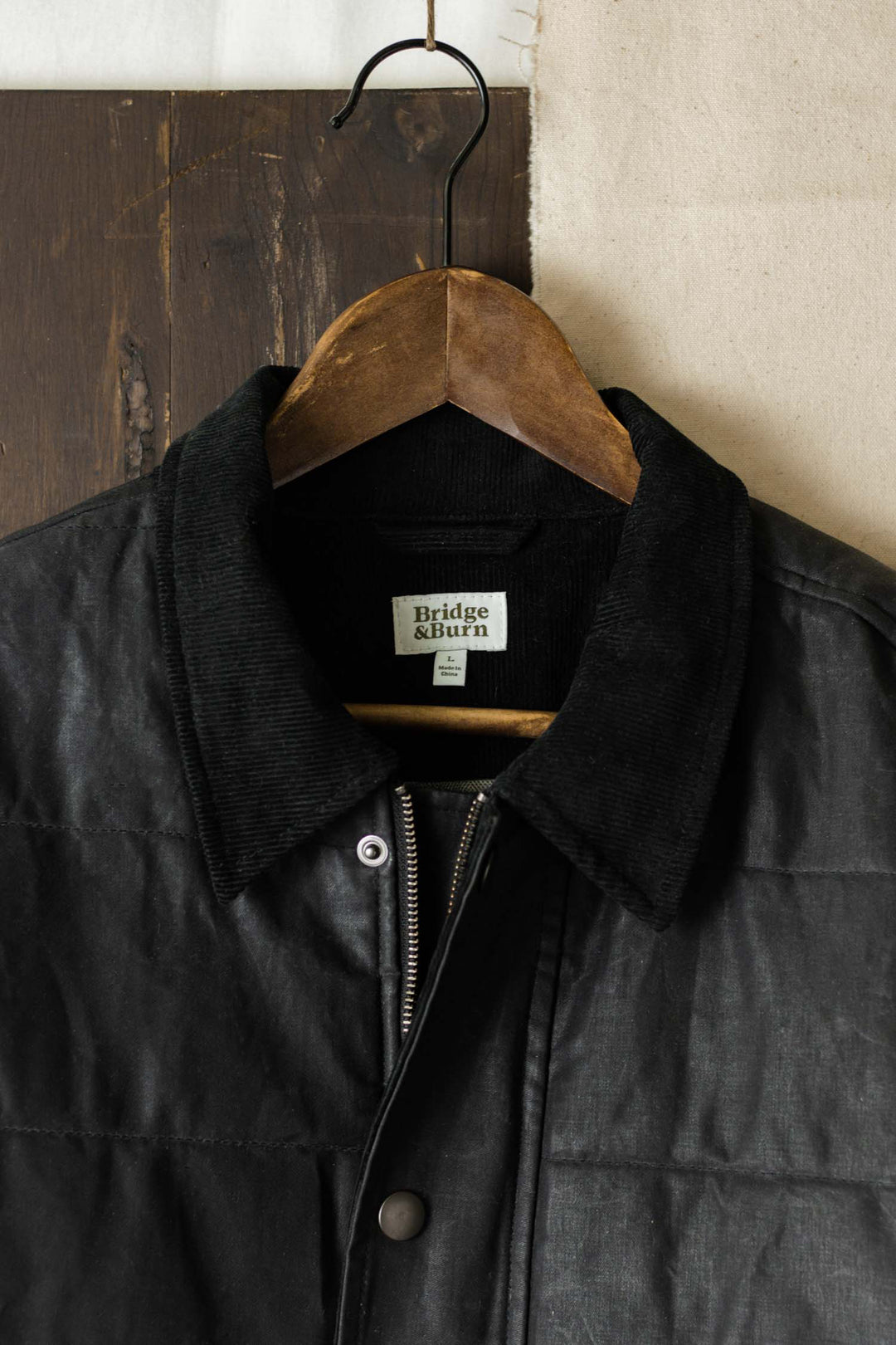 The Holman Quilted Jacket in black waxed canvas by Bridge & Burn hangs on a wooden hanger, showcasing its corduroy collar against a rustic wooden and cream background.