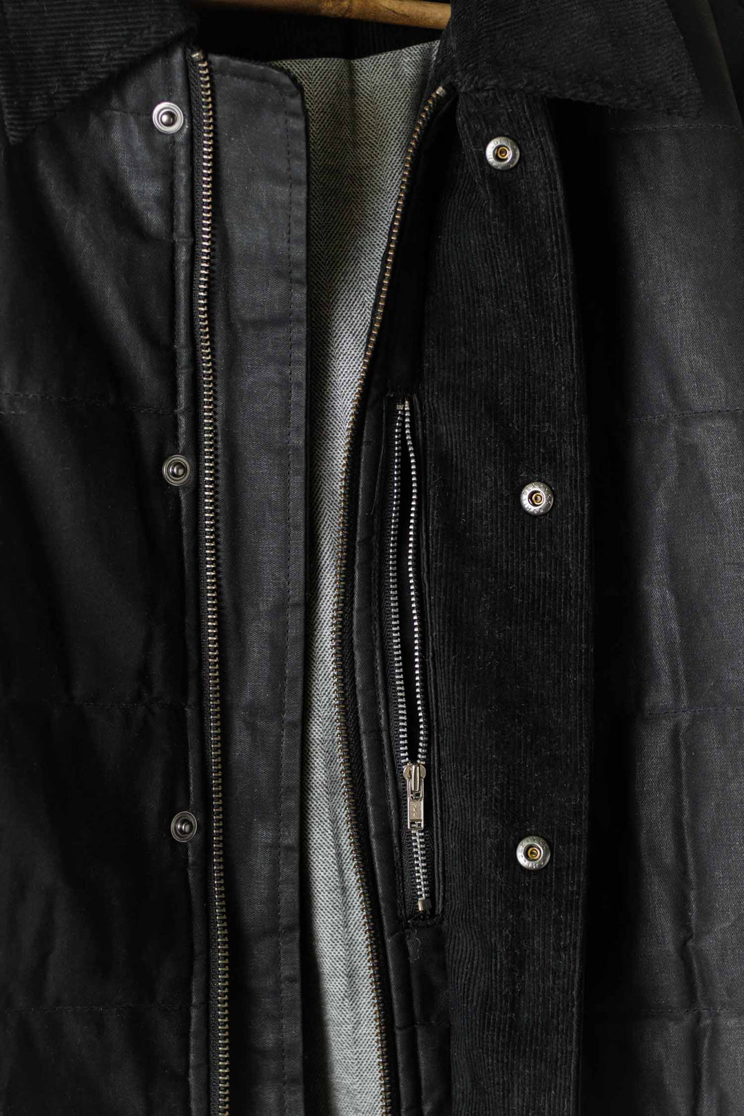 Close-up of the Bridge & Burn Holman Quilted Jacket in black waxed canvas, featuring metal snap buttons, a partially unzipped front showing the light gray lining, and a textured outer finish.