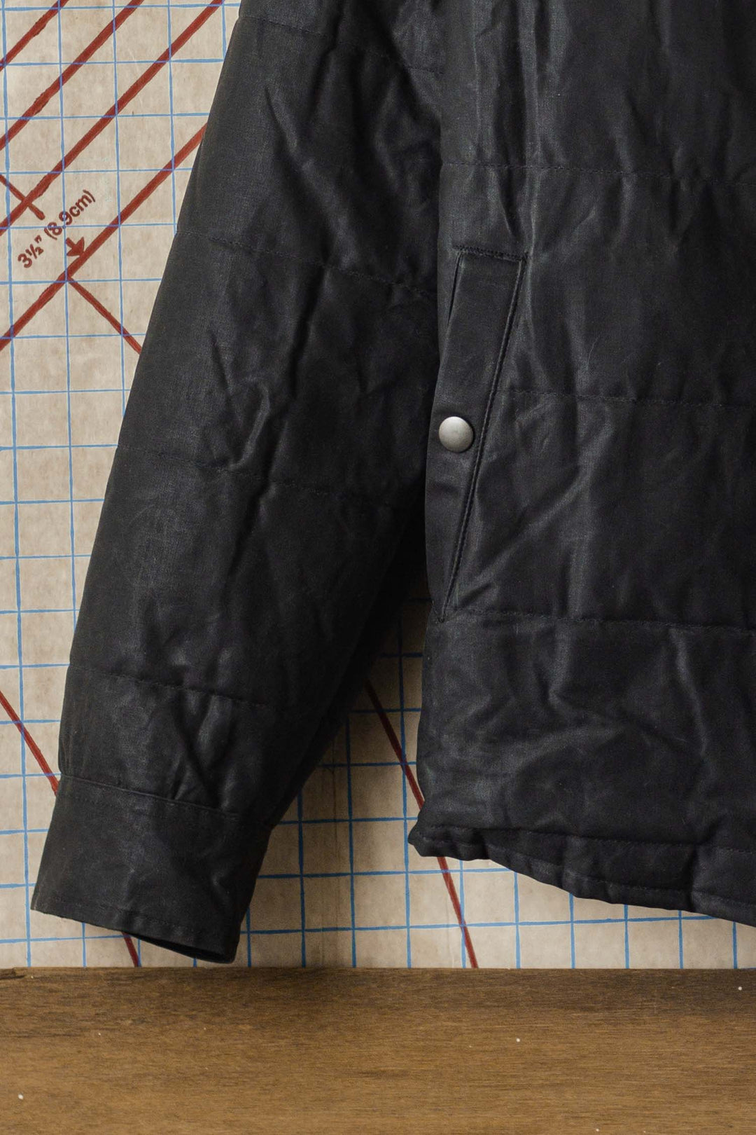 A close-up of the lower sleeve and side of the Bridge & Burn Holman Quilted Jacket in black waxed canvas, featuring a silver snap button, set against a grid-lined background with red markings and a wooden surface beneath.