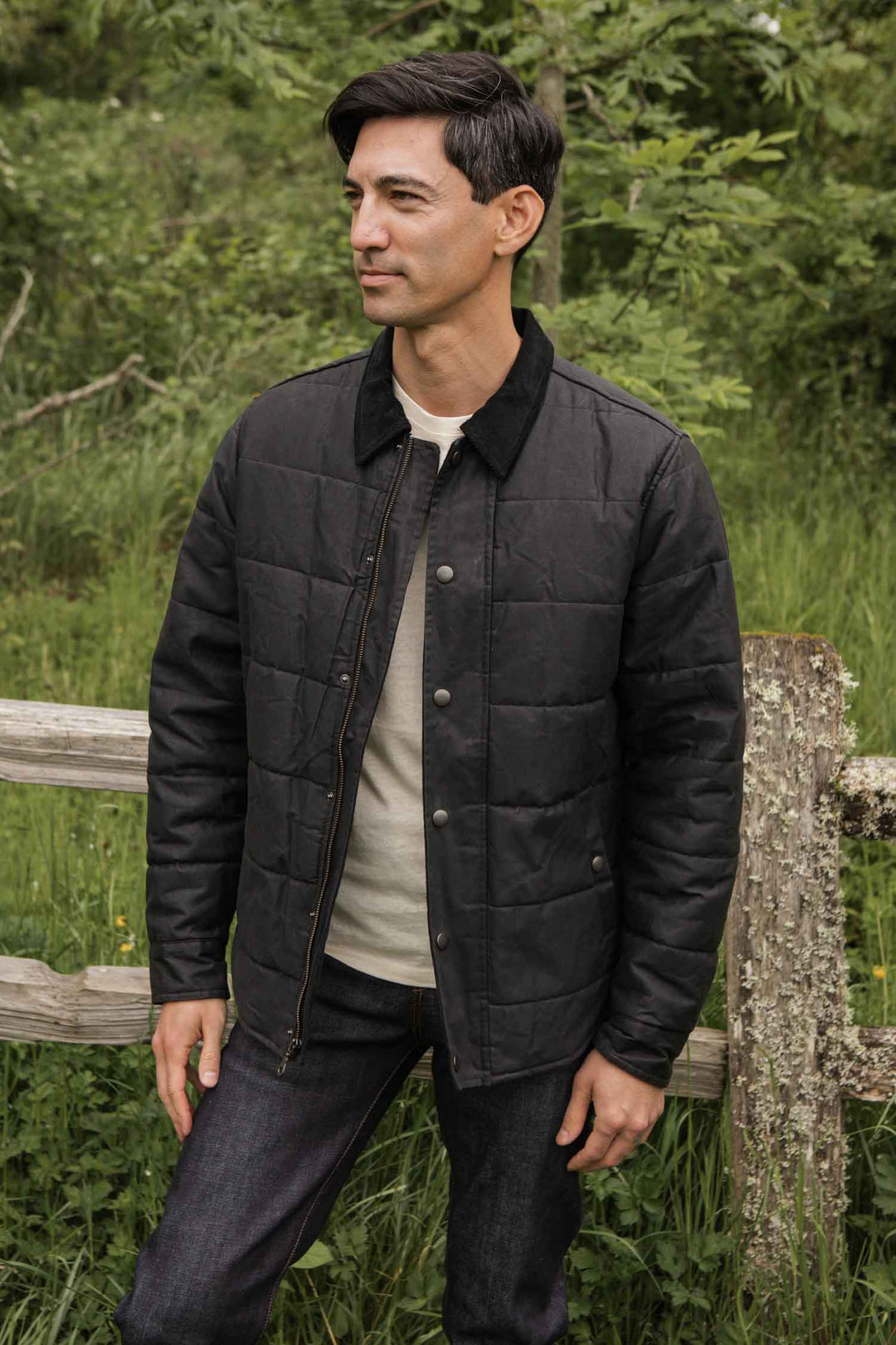 A man stands outdoors in front of a wooden fence and greenery, wearing the Bridge & Burn Holman Quilted Jacket in black waxed canvas over a light shirt and dark jeans. He looks slightly to the side with a neutral expression.