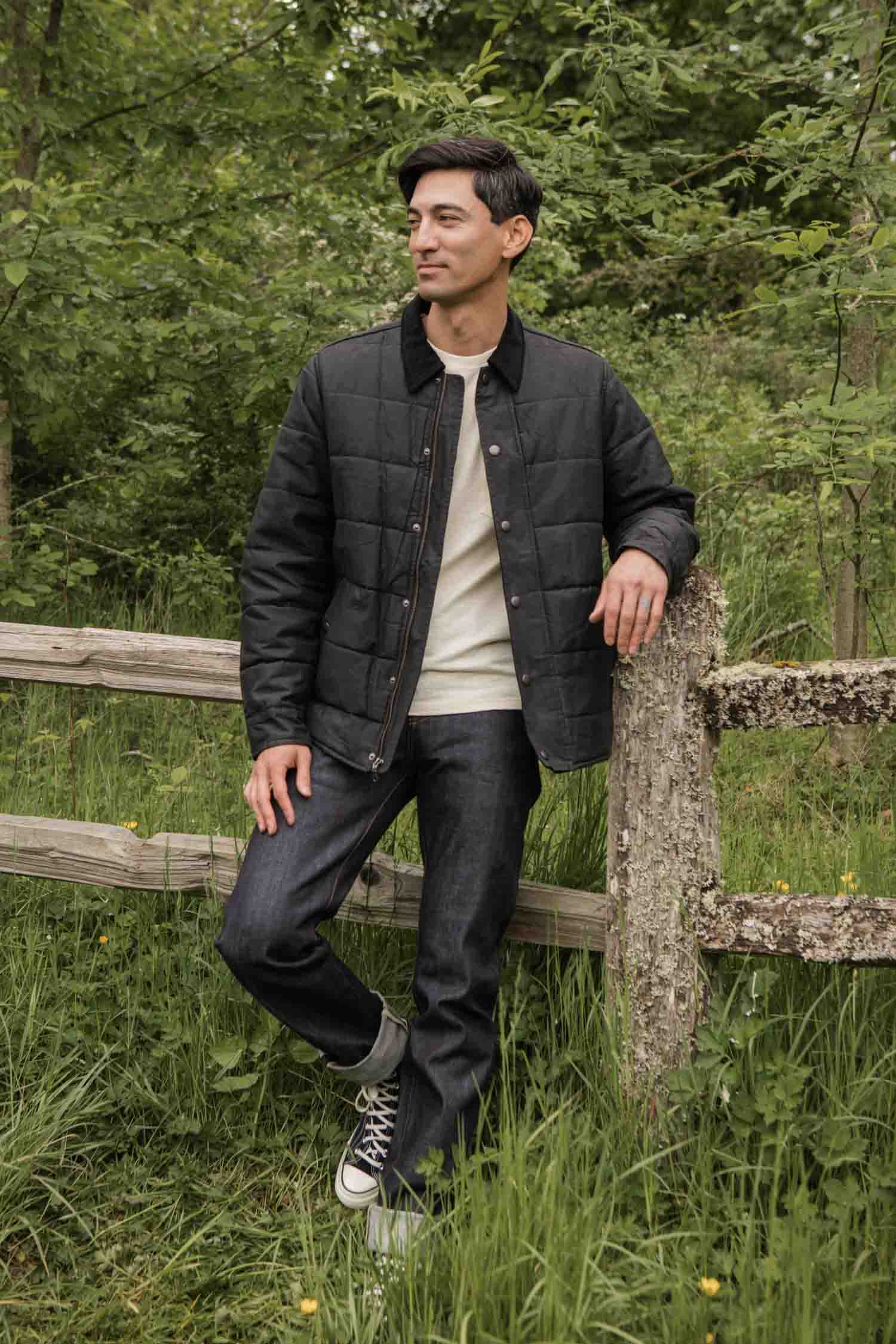 A man in a Bridge & Burn Holman Quilted Jacket in black waxed canvas, a cream sweater, dark jeans, and black sneakers stands by a wooden fence amid tall grass and trees, smiling as he looks off to the side.
