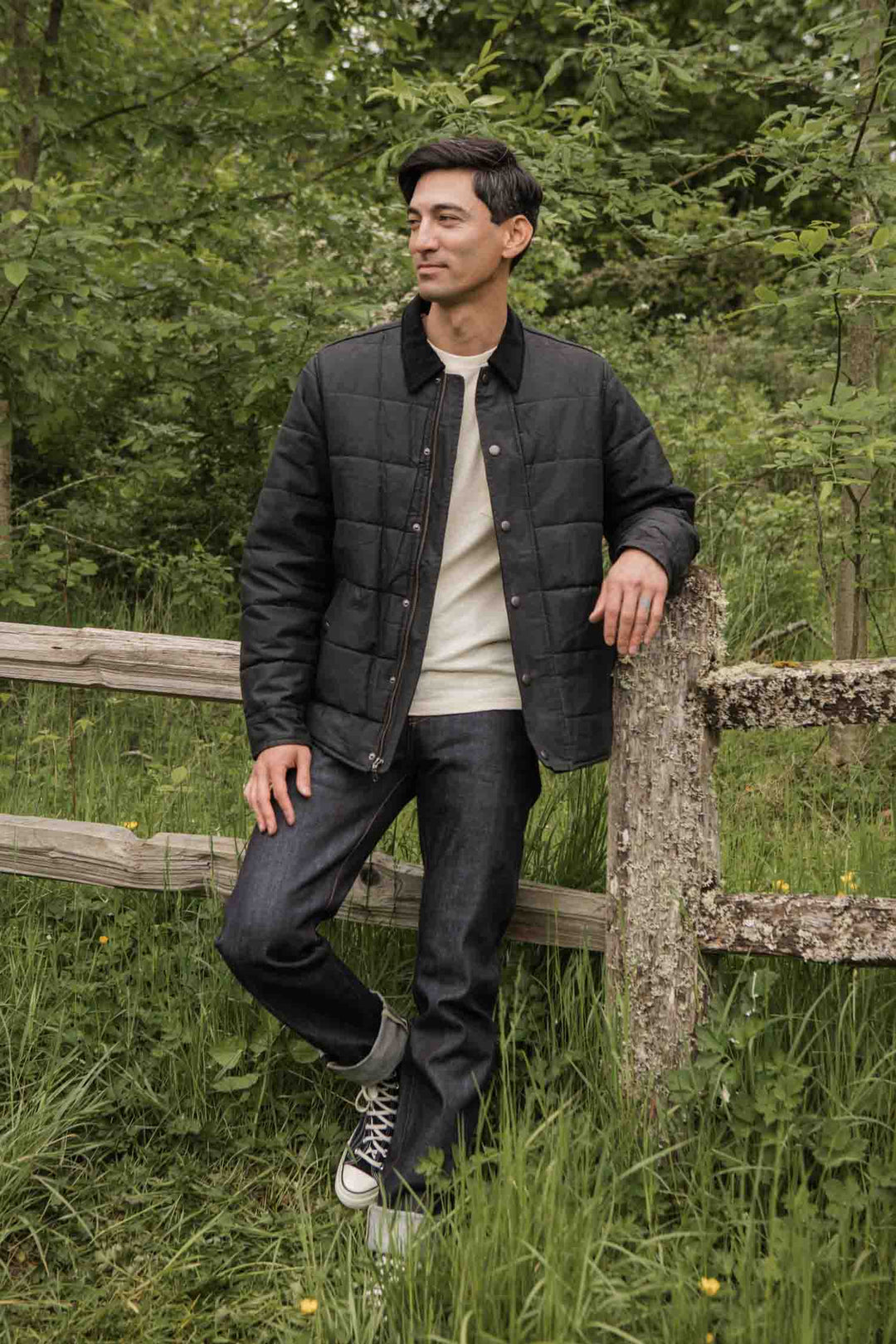 A man in a Bridge & Burn Holman Quilted Jacket in black waxed canvas, a cream sweater, dark jeans, and black sneakers stands by a wooden fence amid tall grass and trees, smiling as he looks off to the side.