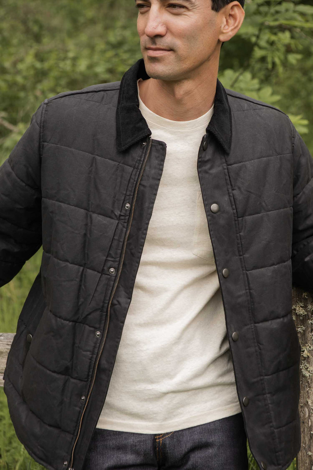 A man stands outdoors by wooden fencing and greenery, wearing the Bridge & Burn Holman Quilted Jacket in black waxed canvas over a white shirt.